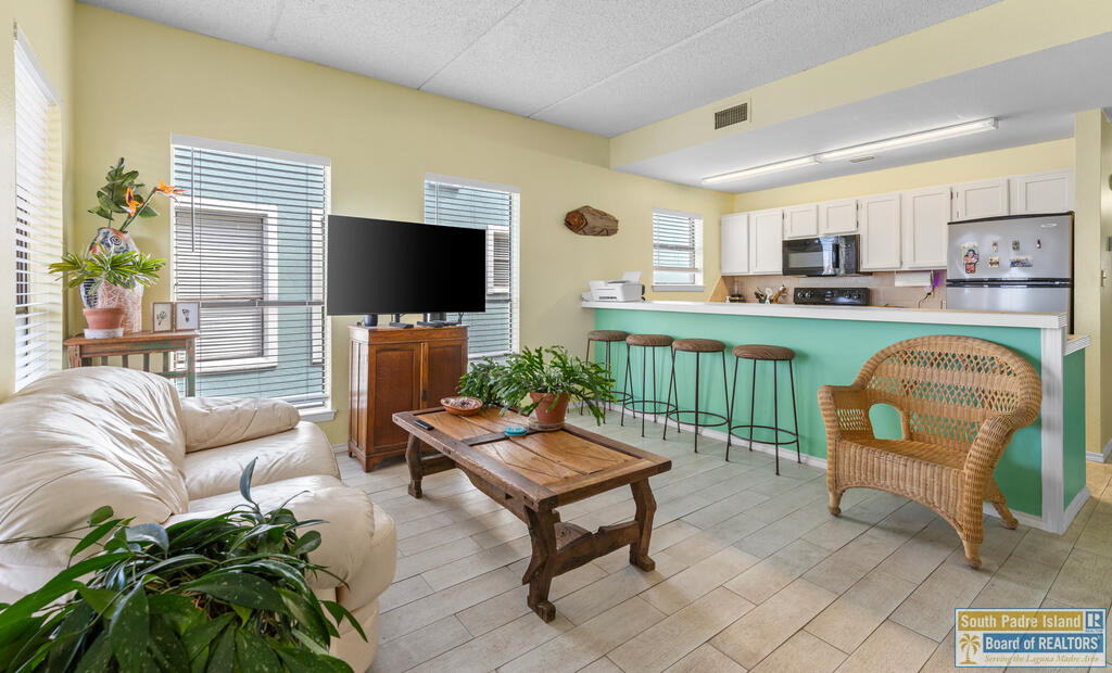 Living room with a turquoise breakfast bar, wicker armchair, white sofa, and large windows.