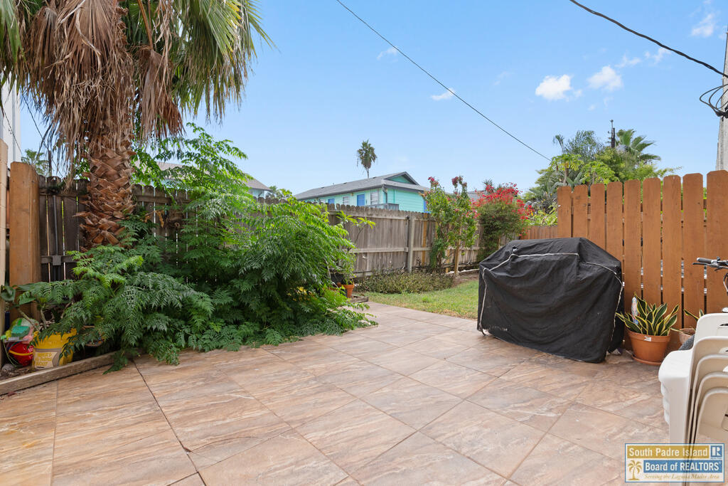 Fenced patio with stone-look pavers, tropical plants, and a covered grill.