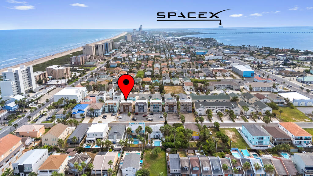 Aerial view of coastal neighborhood marked by a red pin, with beach and ocean beyond.