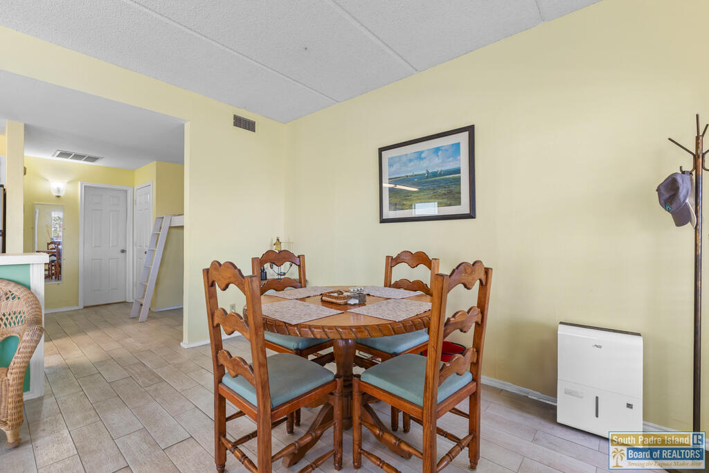 Dining area with a wooden table and six chairs, tiled floors, and a framed coastal landscape.