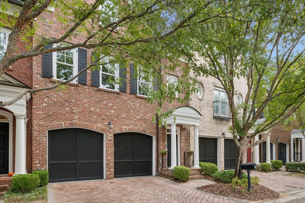 Brick townhouse with black garage doors, white entry columns, and a tree-shaded paver driveway.