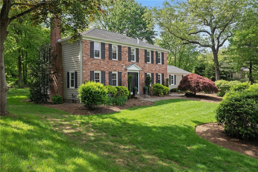 Two-story brick Colonial house with dark shutters, landscaped shrubs, and a broad green lawn.
