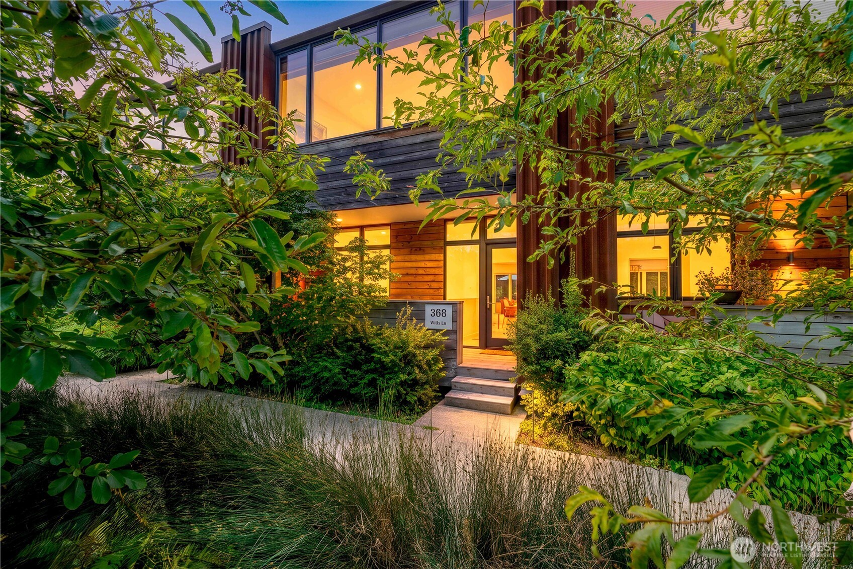 Modern two-story home with glowing windows, dark wood siding, and a lush garden at dusk.