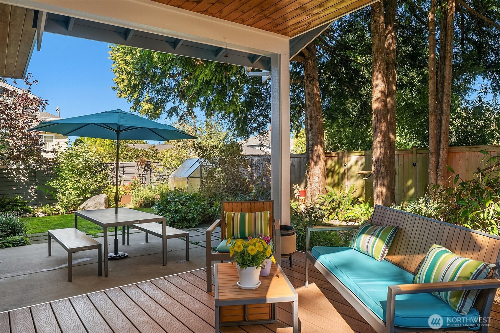 Covered patio with teal-cushioned seating overlooks a leafy garden and tall trees.