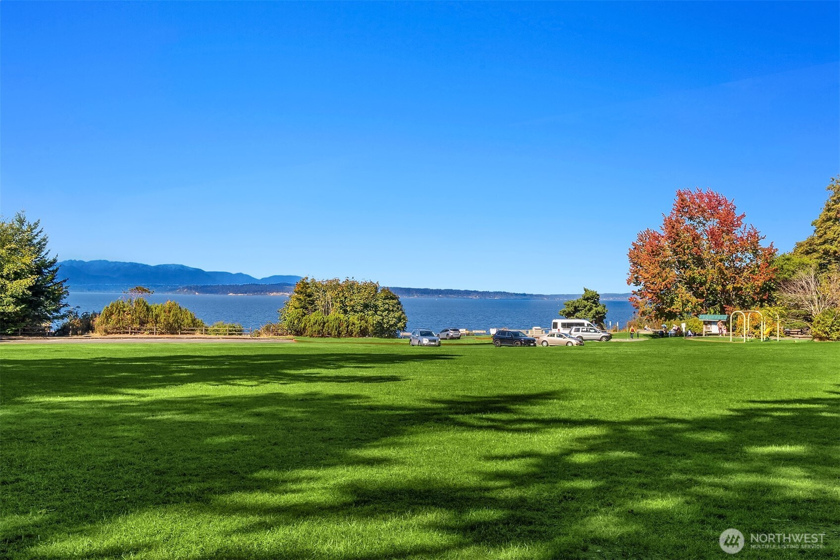 Waterfront park with a wide green lawn, distant mountains, and autumn-colored trees.