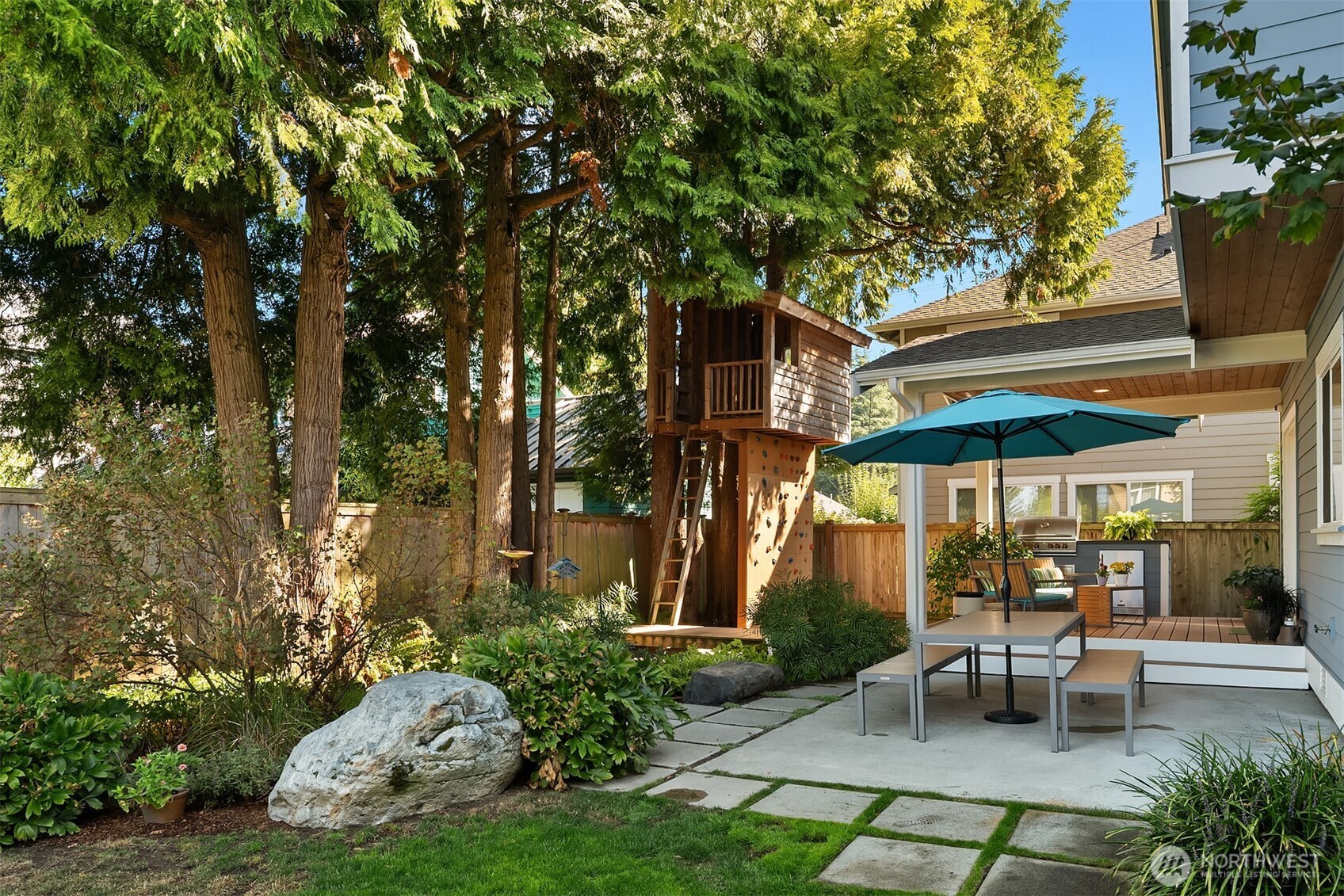 Backyard patio with teal umbrella, landscaped garden, and a wooden treehouse with a climbing wall.