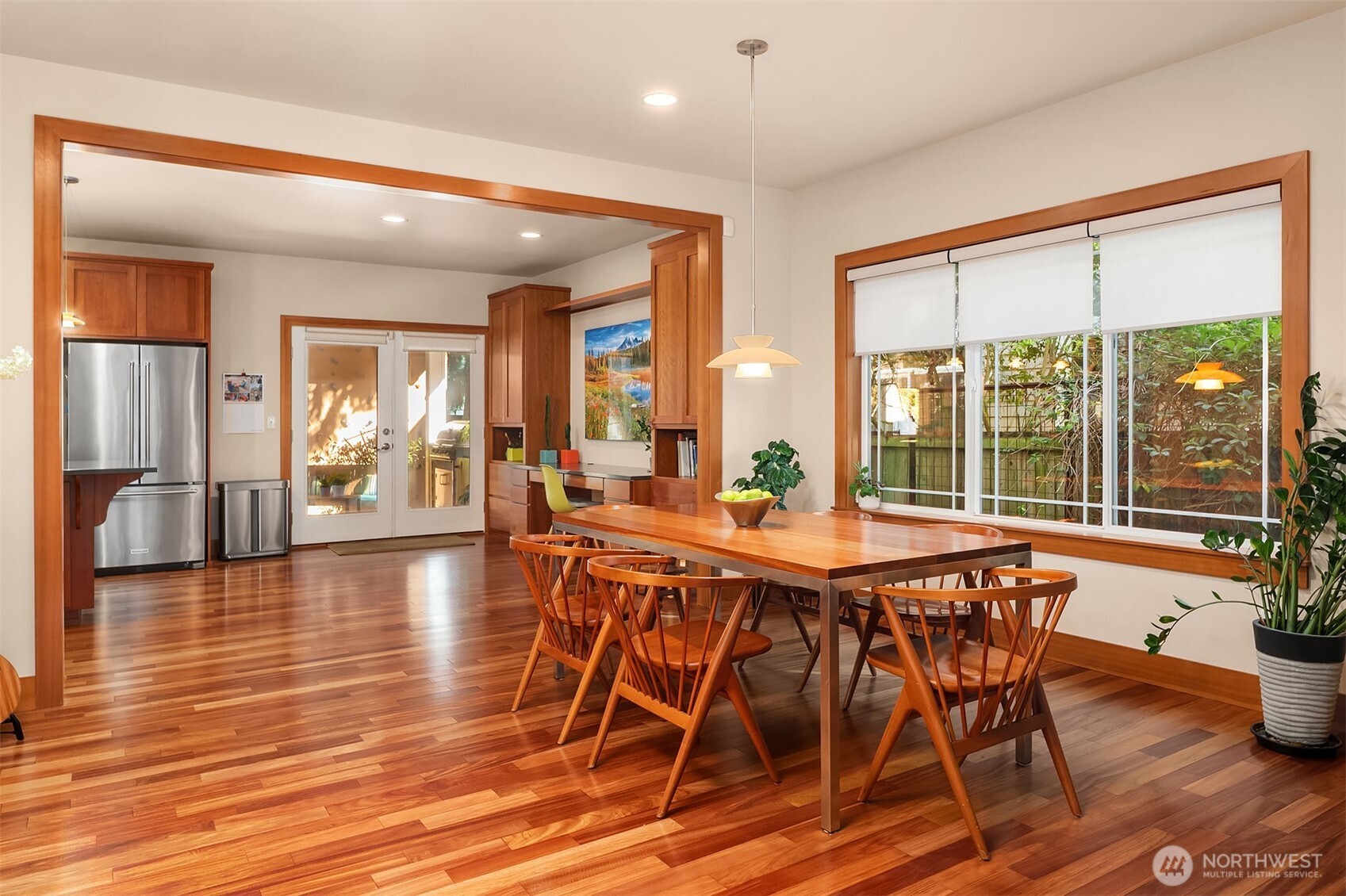 Dining area with a long wooden table, matching chairs, hardwood floors, and a large window overlooking greenery.
