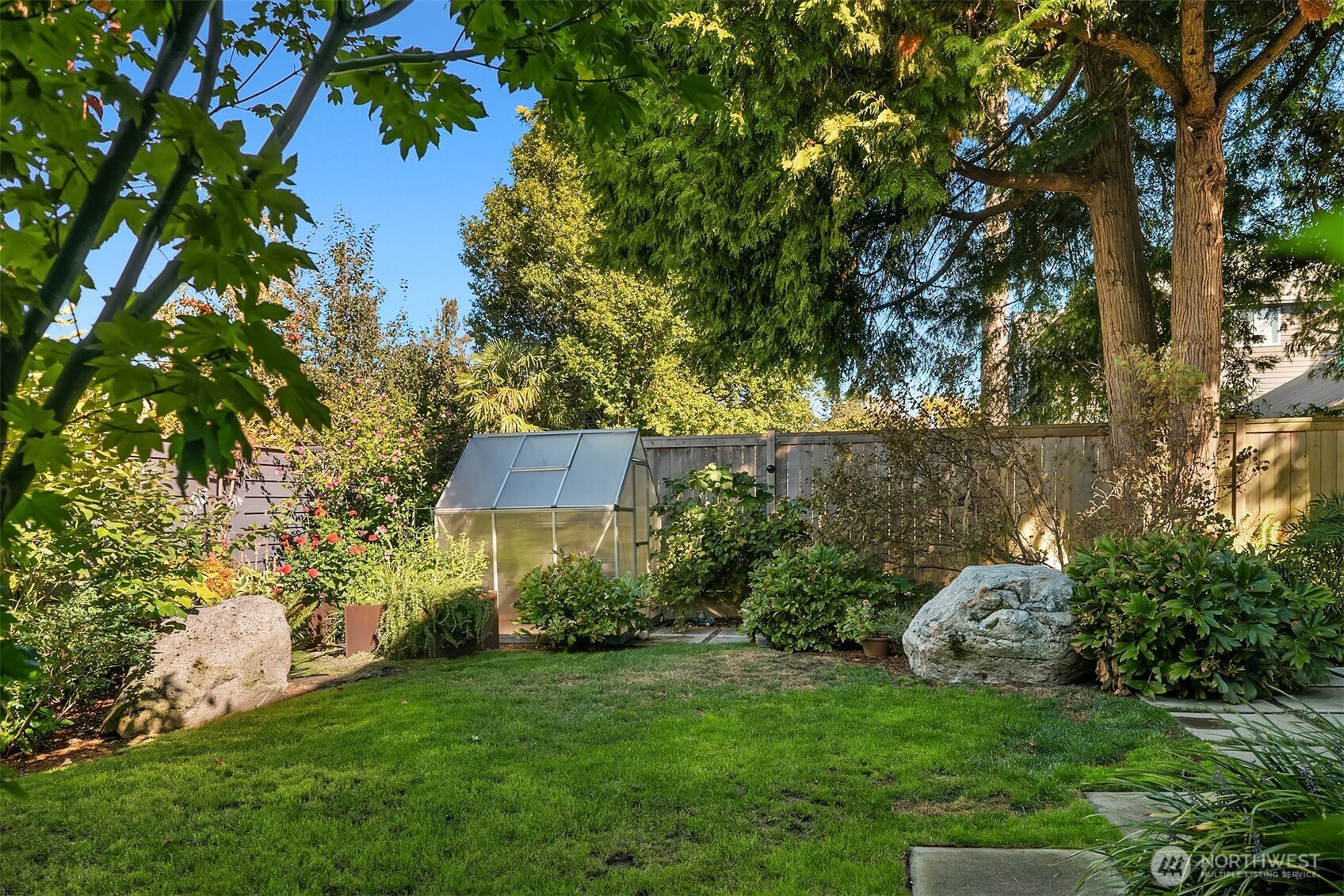 Landscaped backyard with a small greenhouse, flowering plants, mature trees, and a broad lawn.