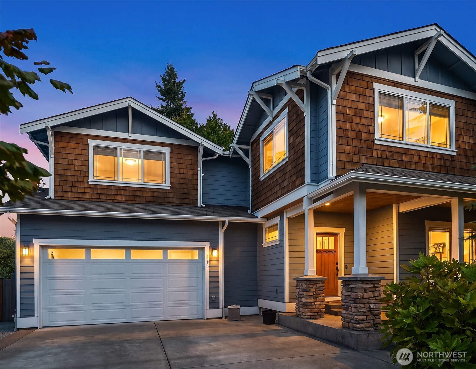 Two-story blue-gray and brown-shingle home with a lit porch and two-car garage at dusk.