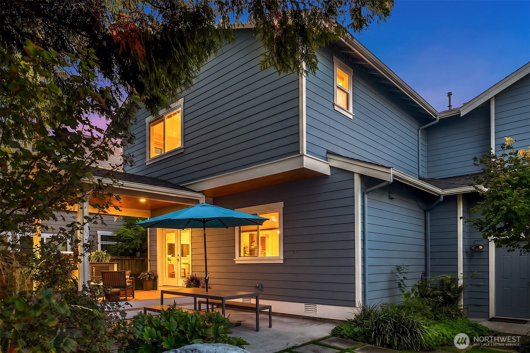 Two-story blue-gray house at dusk with warmly lit windows and a covered patio.