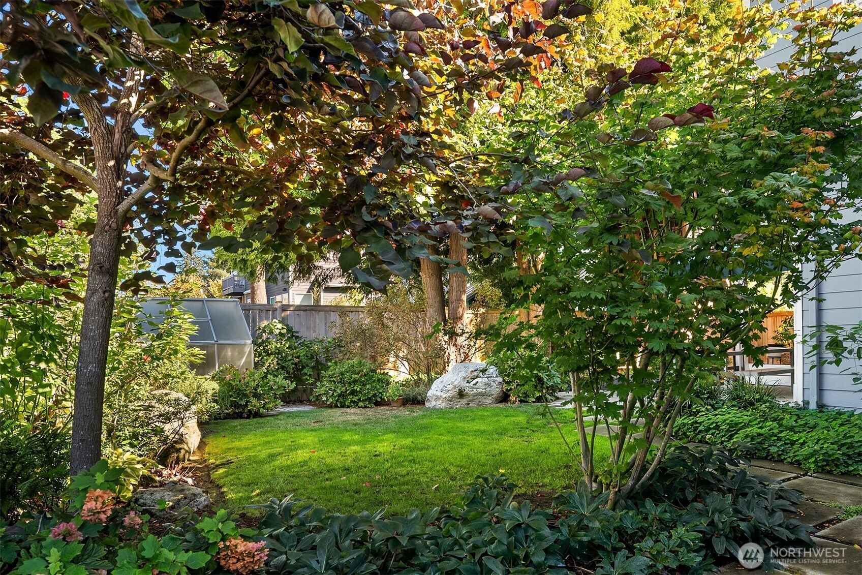 Landscaped backyard with a green lawn, mature autumn trees, large rocks, and a small greenhouse.
