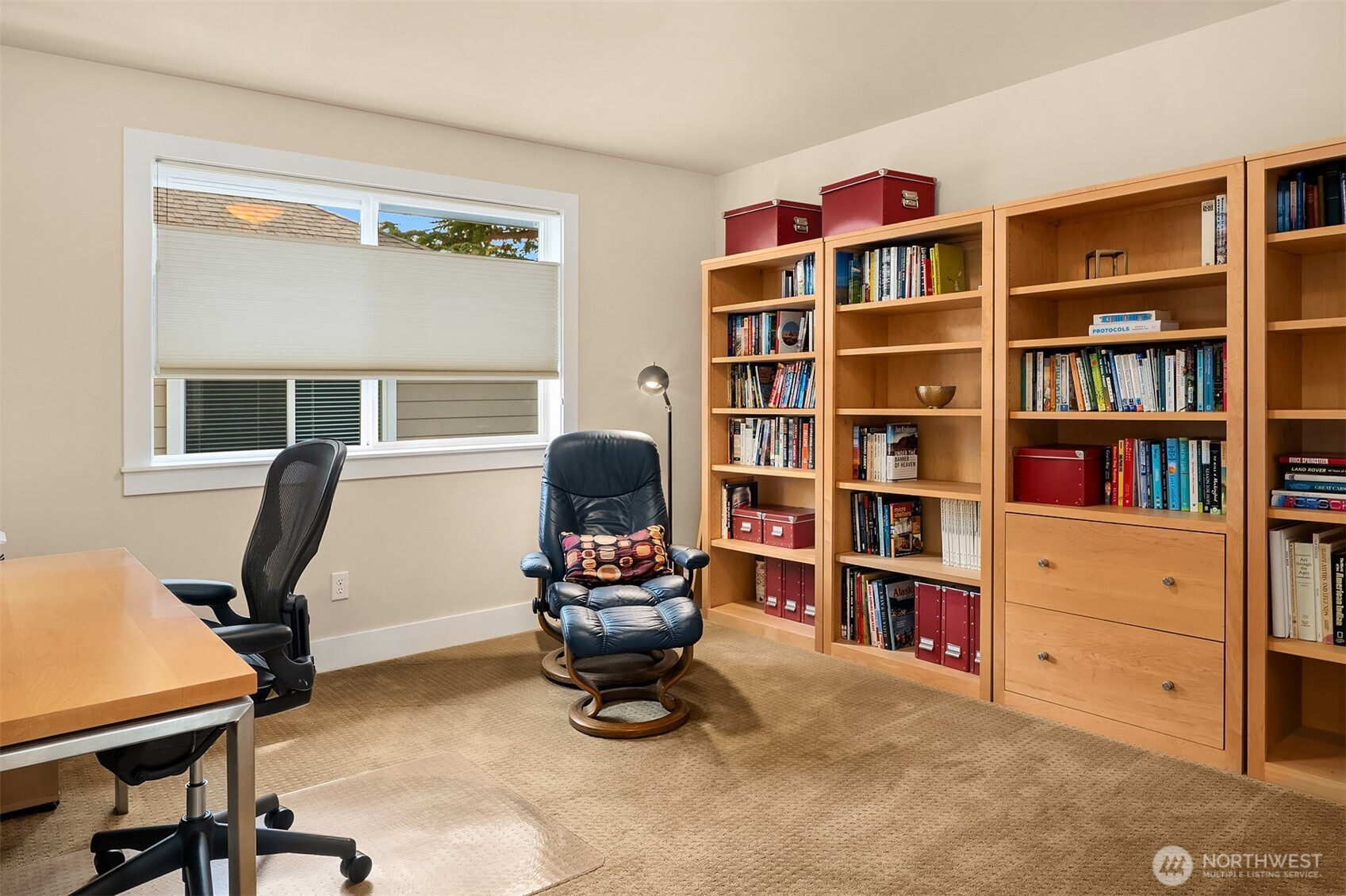 Home office with wooden bookcases, a desk, and a leather lounge chair beside a large window.