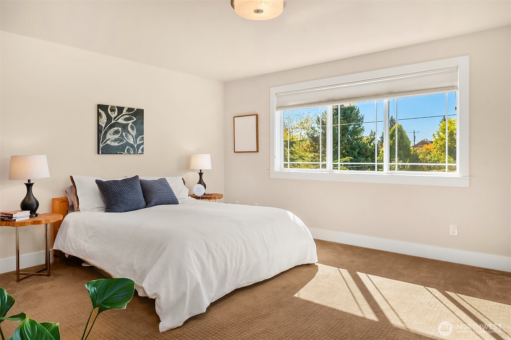Bedroom with a white bed, dark pillows, and a wide sunny window overlooking trees.