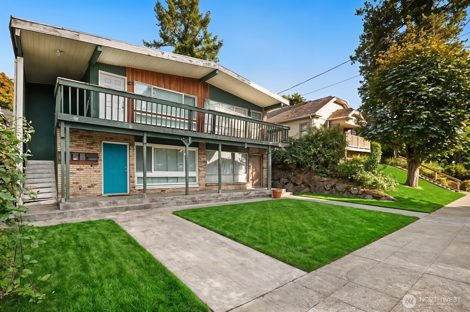 Two-story home with a long balcony, green and wood siding, and a bright blue front door.