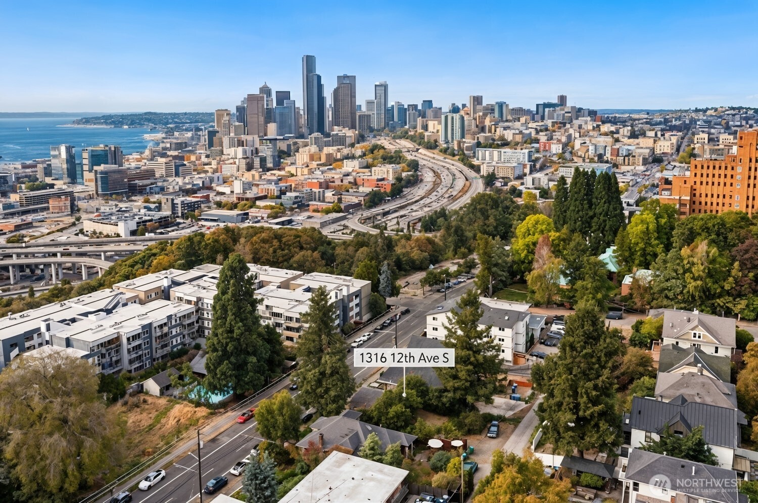 Aerial view of 1316 12th Avenue South, with downtown Seattle’s skyline, Puget Sound, and tree-lined streets beyond.