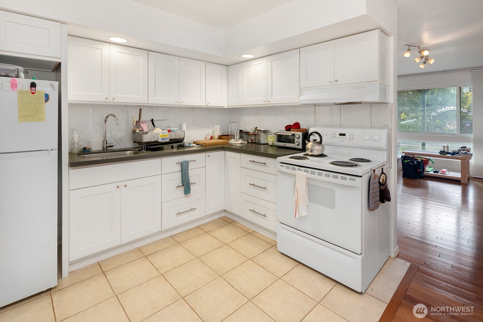 Kitchen with white cabinets, dark counters, and beige tile flooring, opening to a bright room with large windows.