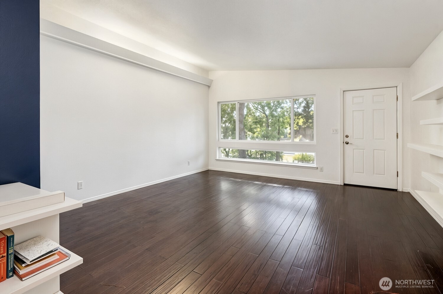 Vacant room with dark wood floors, a large tree-facing window, and built-in white shelving.