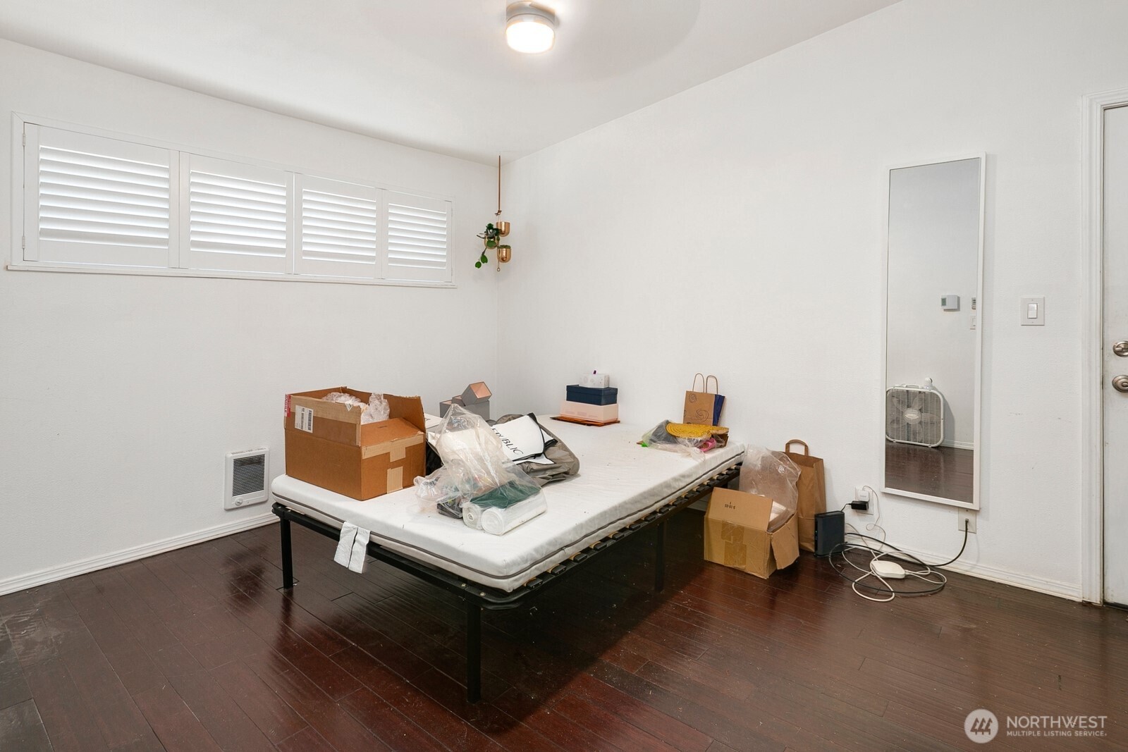Bedroom with dark wood floors, shuttered windows, and a mattress surrounded by boxes and belongings.