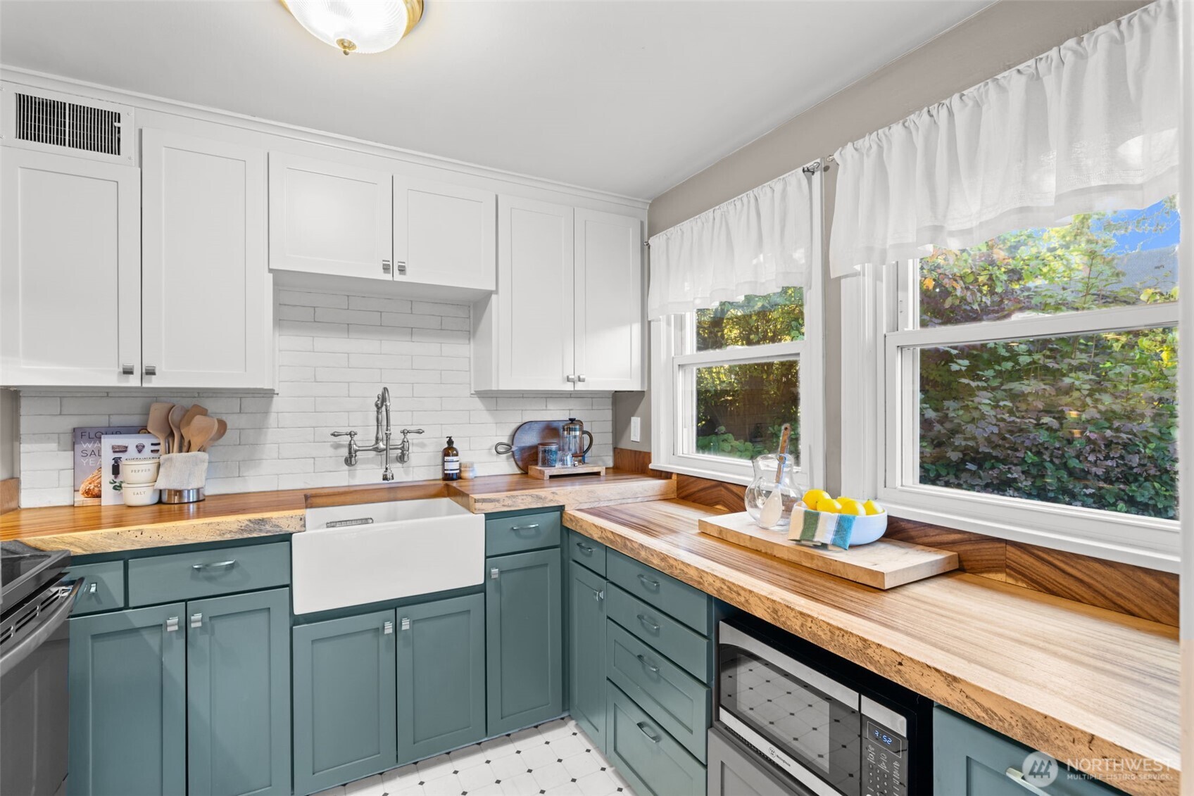 Kitchen with blue-green cabinets, butcher-block counters, farmhouse sink, and windows overlooking greenery.