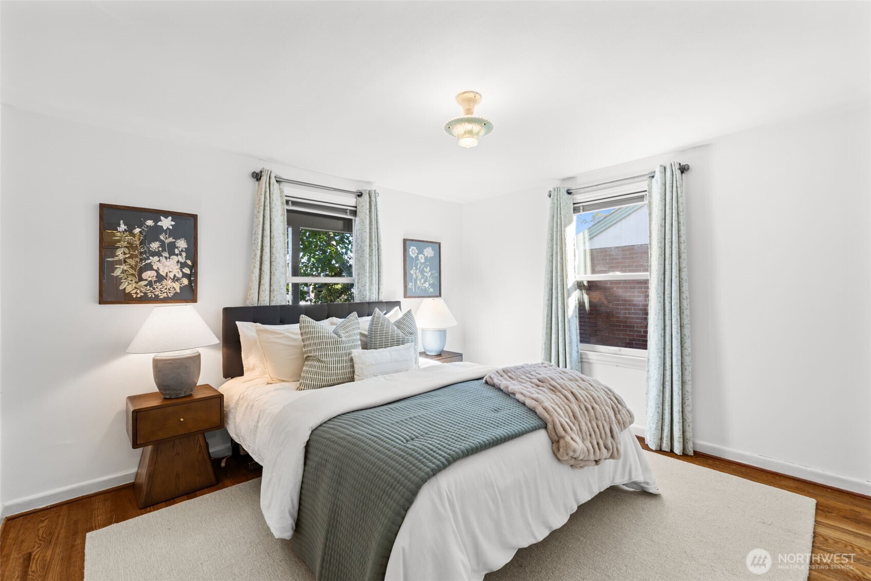 Staged bedroom with a large bed, botanical prints, and pale curtains framing two windows.