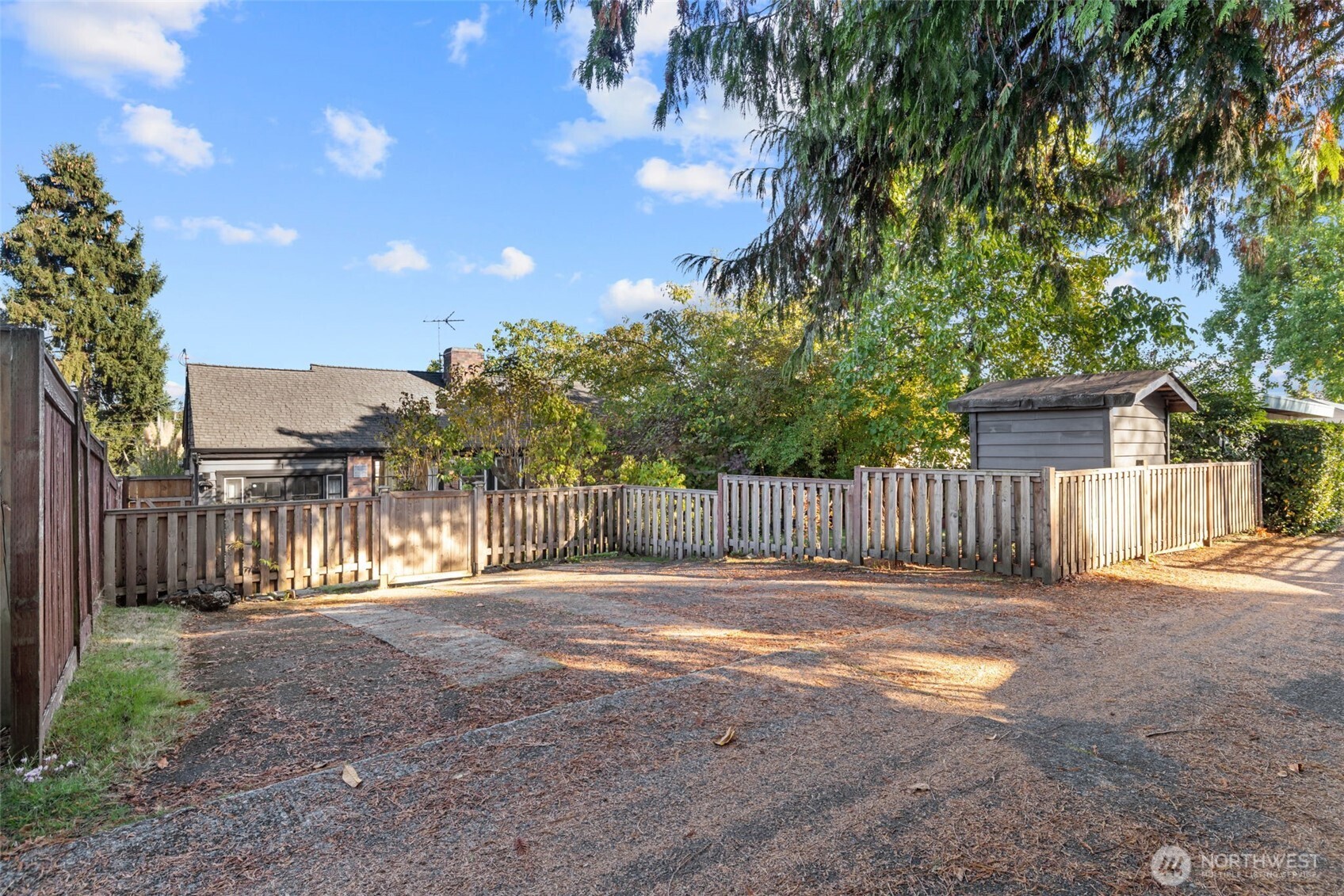 Fenced backyard with a broad paved area, mature trees, and a small wooden shed.