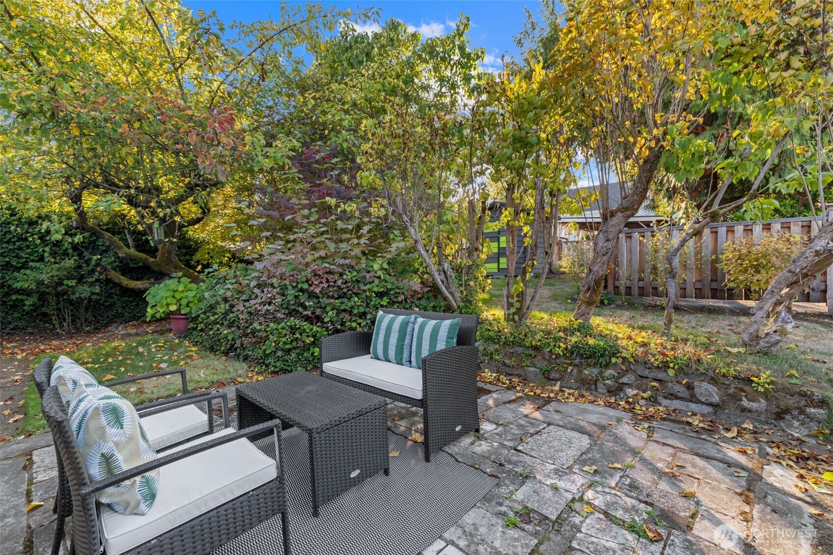 Backyard patio with wicker seating and a stone-paver terrace beneath mature autumn trees.