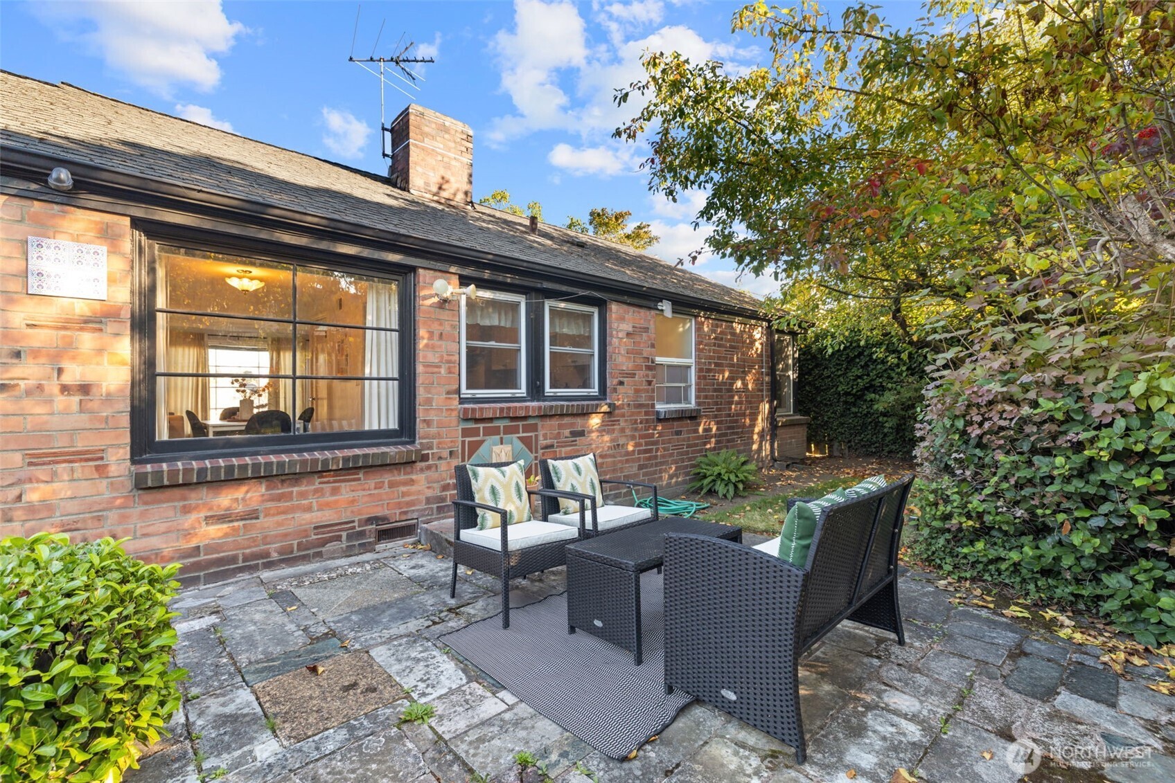 Stone patio with wicker seating beside a brick house, surrounded by leafy hedges and trees.
