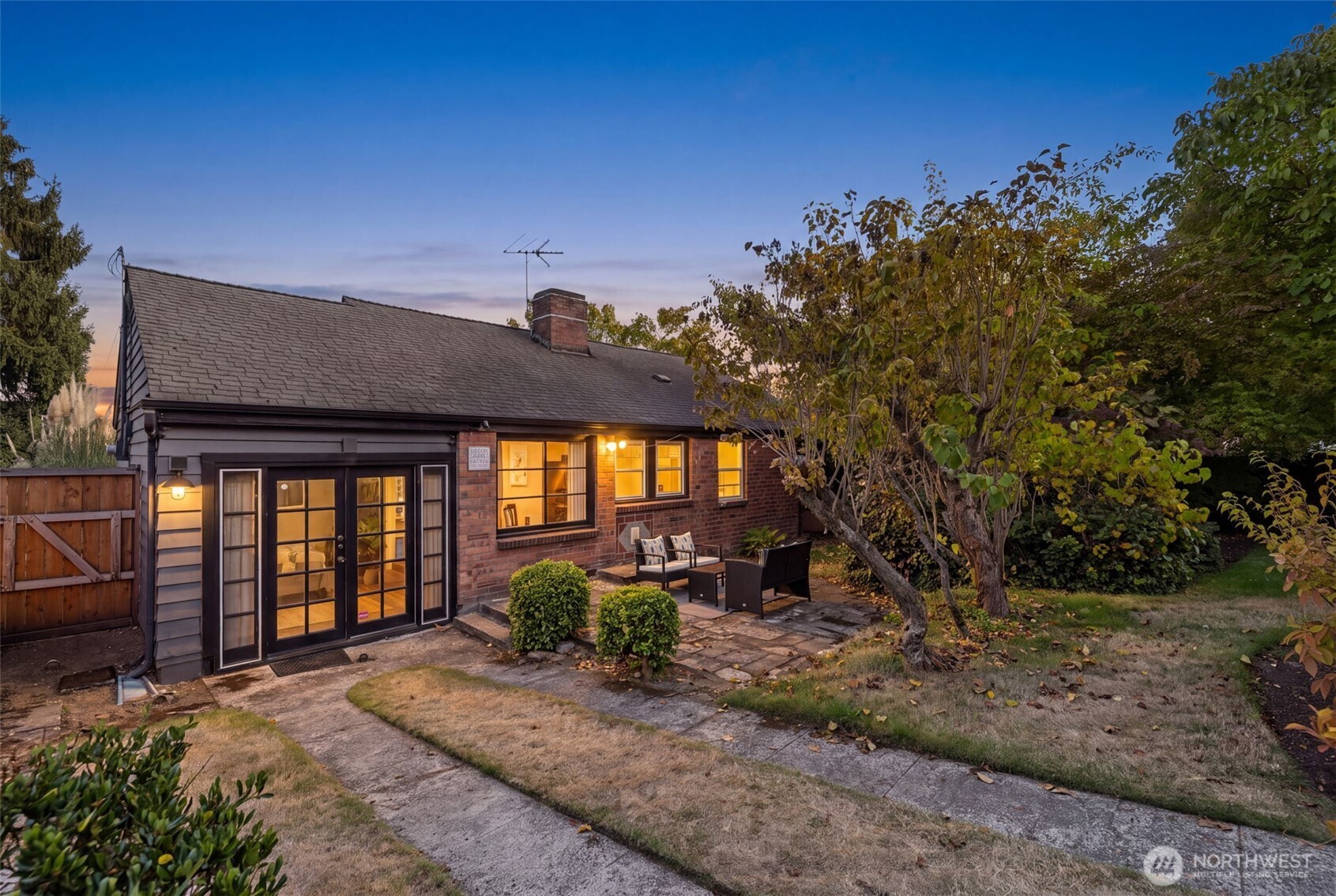 Single-story brick home glowing at dusk, with a stone patio and autumn trees.