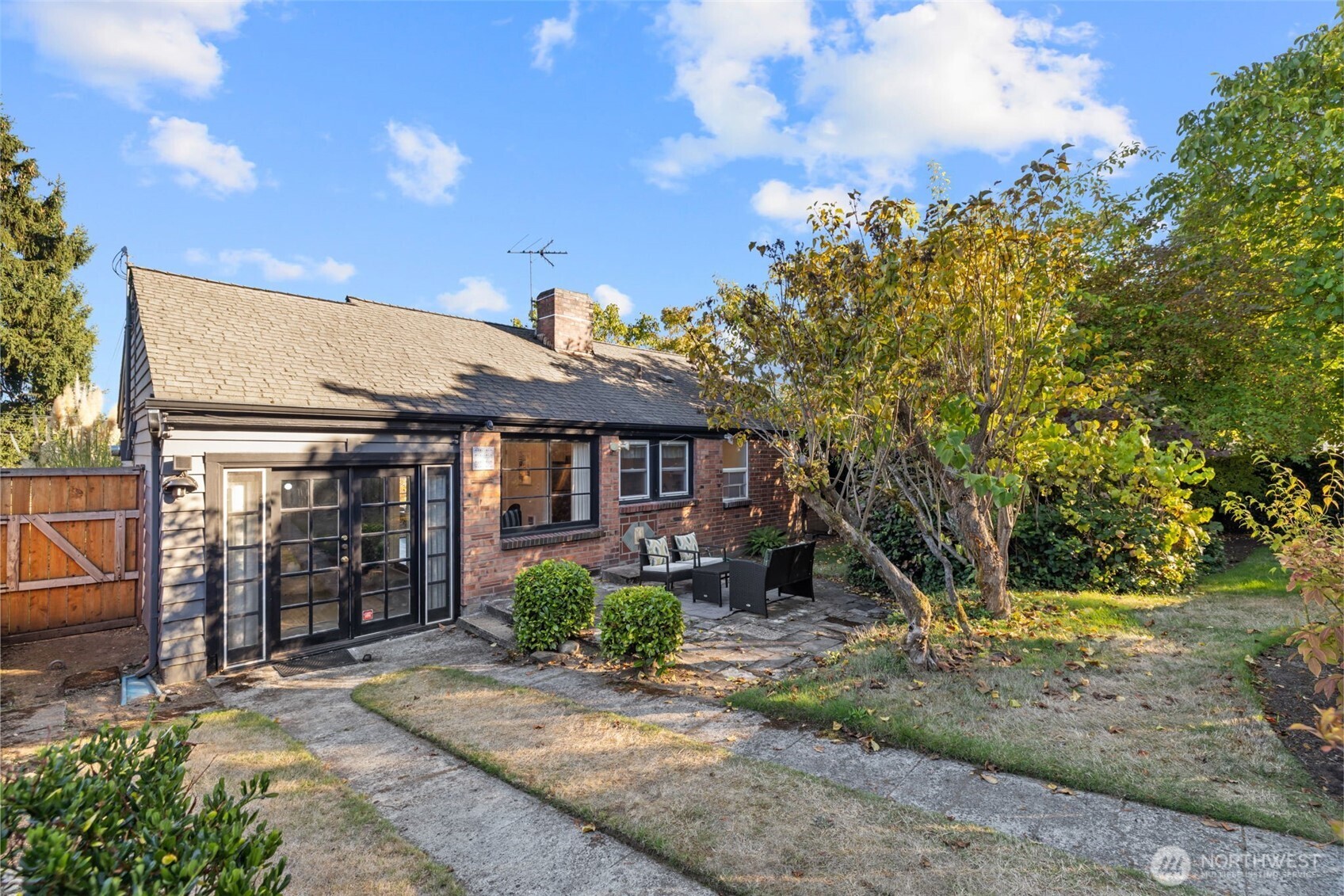 Brick-and-siding house with a stone patio, outdoor seating, and mature garden trees.