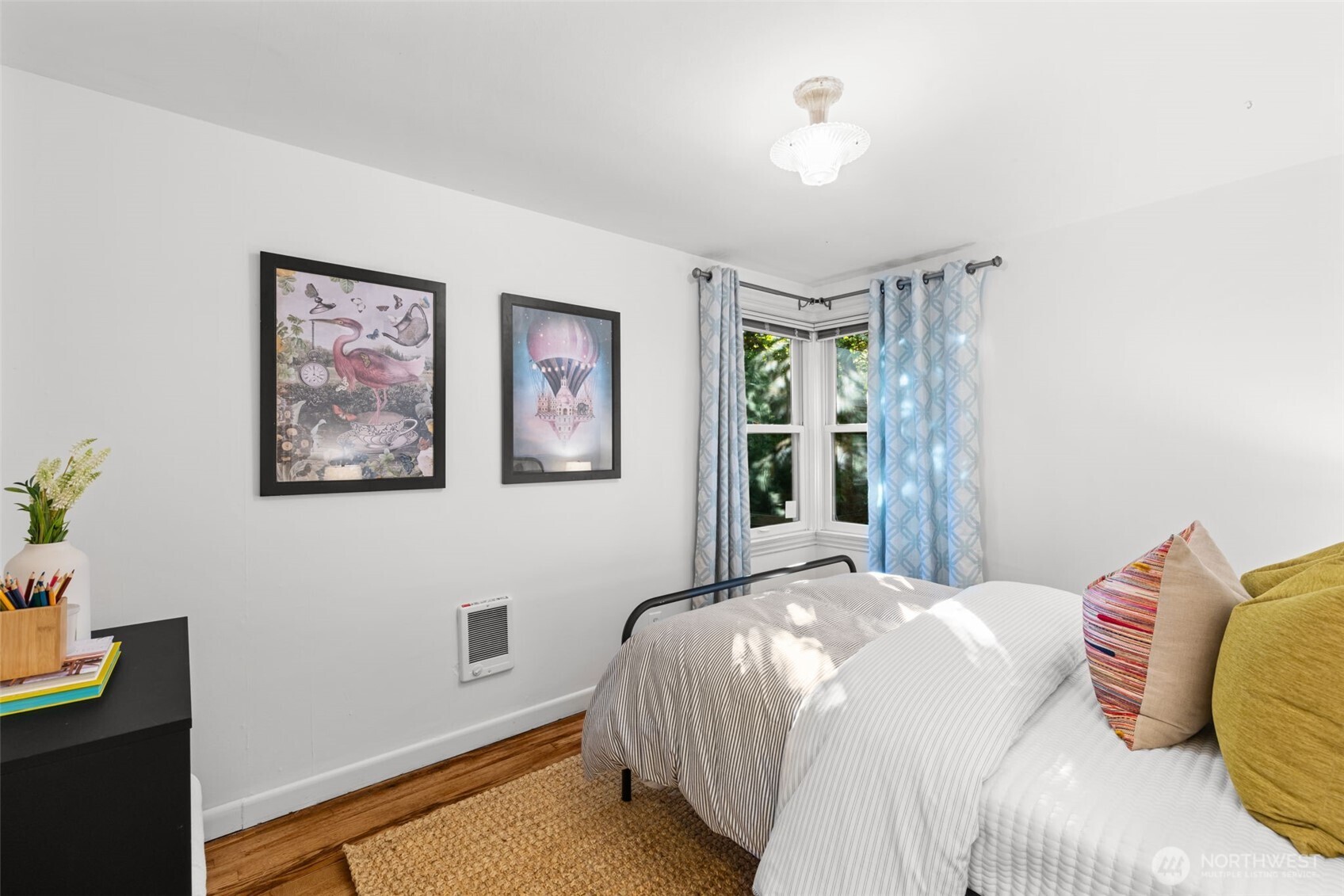 Furnished bedroom with striped bedding, colorful pillows, framed art, and a corner window with blue curtains.