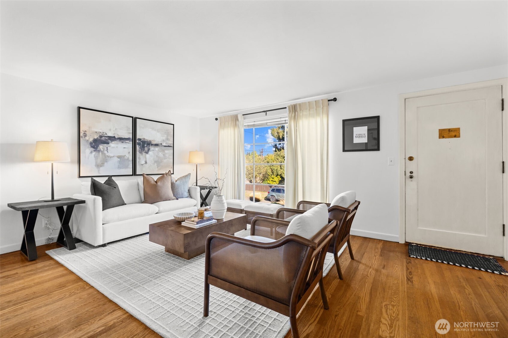 Staged living room with a white sofa, woven chairs, hardwood floors, and a large curtained window.