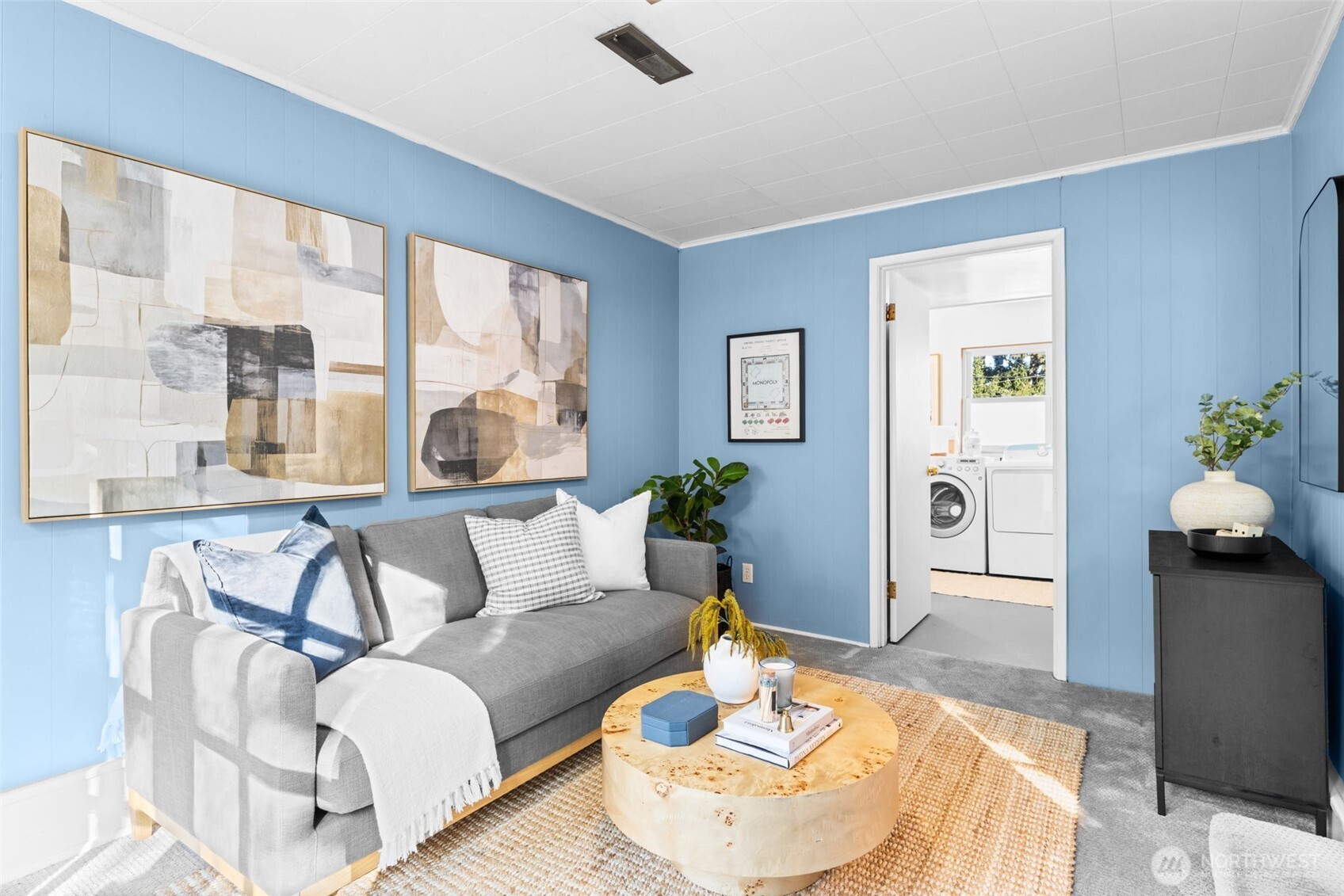 Staged living room with blue paneled walls, gray sofa, abstract artwork, and doorway to a laundry room.