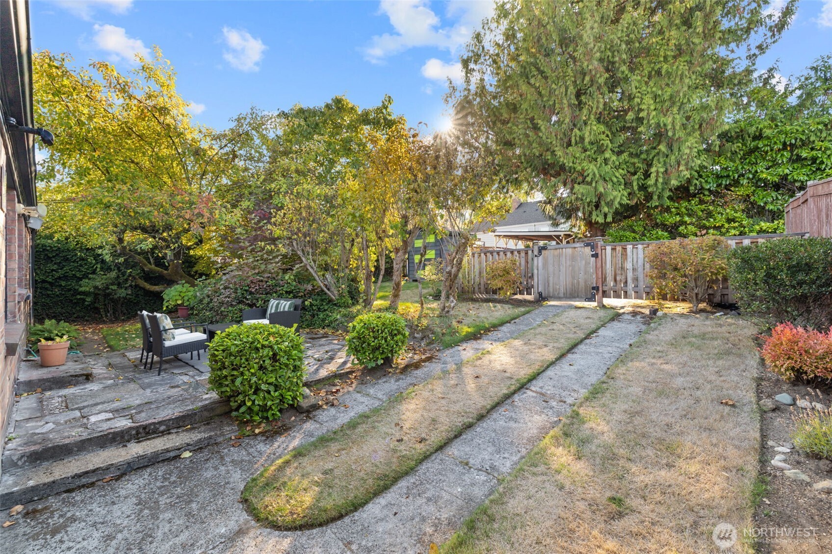 Backyard patio with outdoor seating, mature trees, and concrete paths leading to a wooden gate.