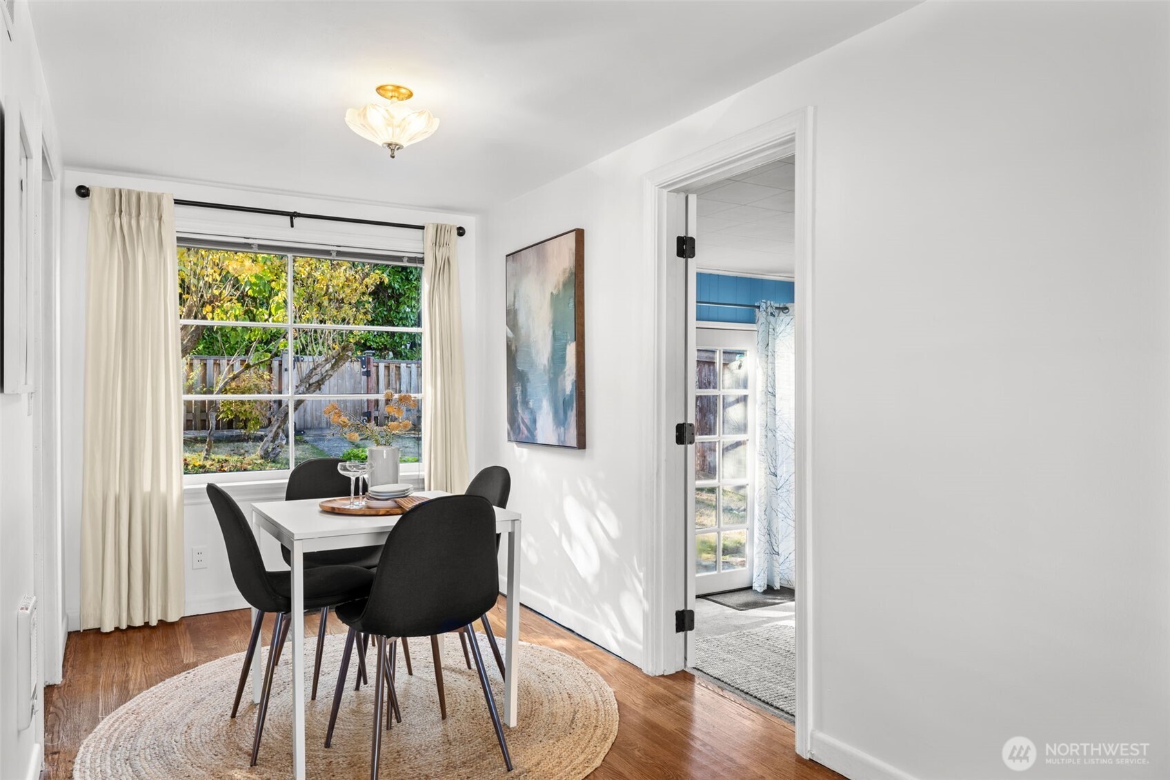 Dining room with a white table, black chairs, hardwood floors, and a large window overlooking a fenced yard.
