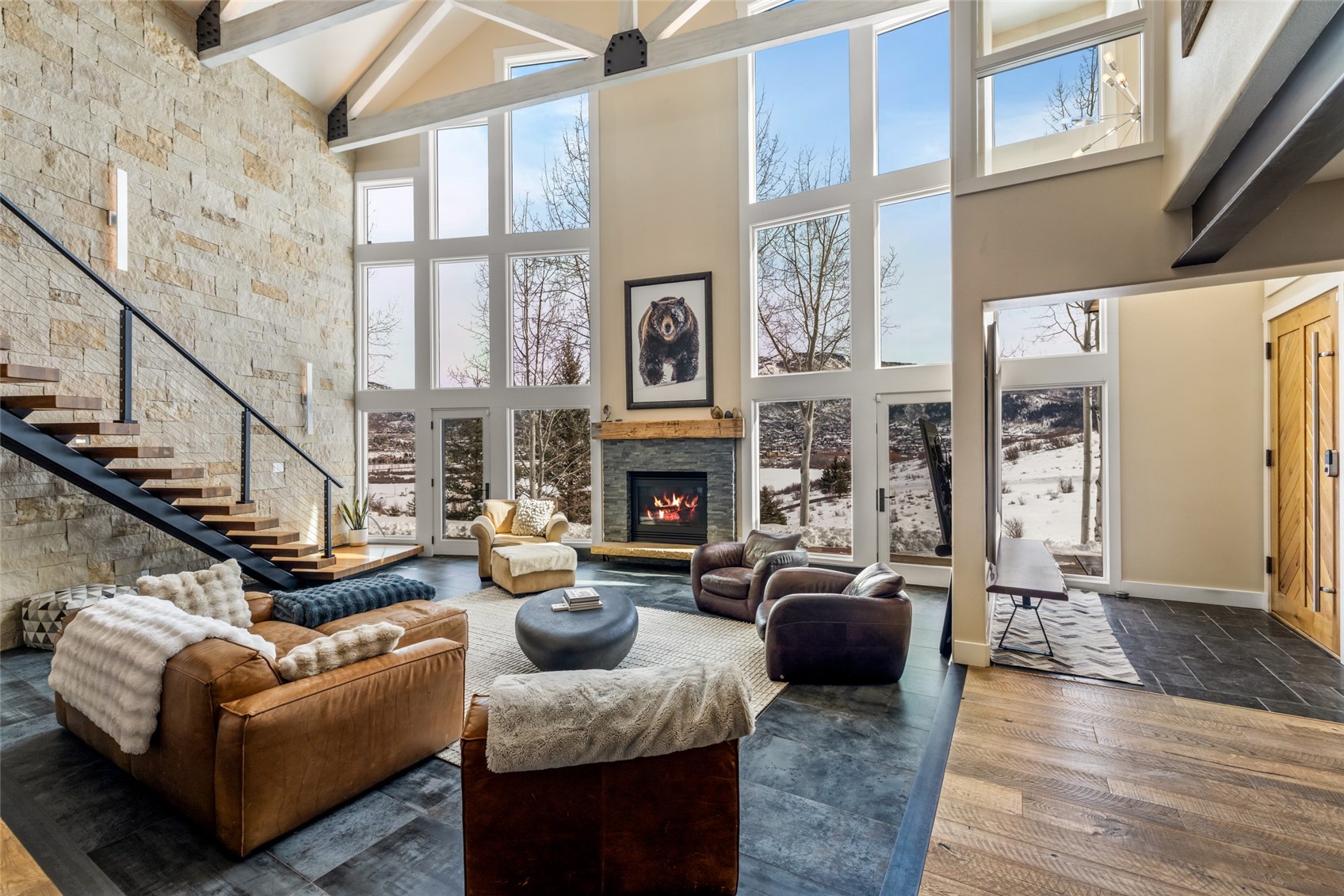 Double-height living room with a stone fireplace, floating staircase, and snowy mountain views through tall windows.