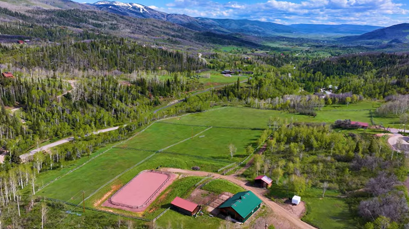 Aerial view of green pastures, barns, and a riding arena beneath snow-capped mountains.