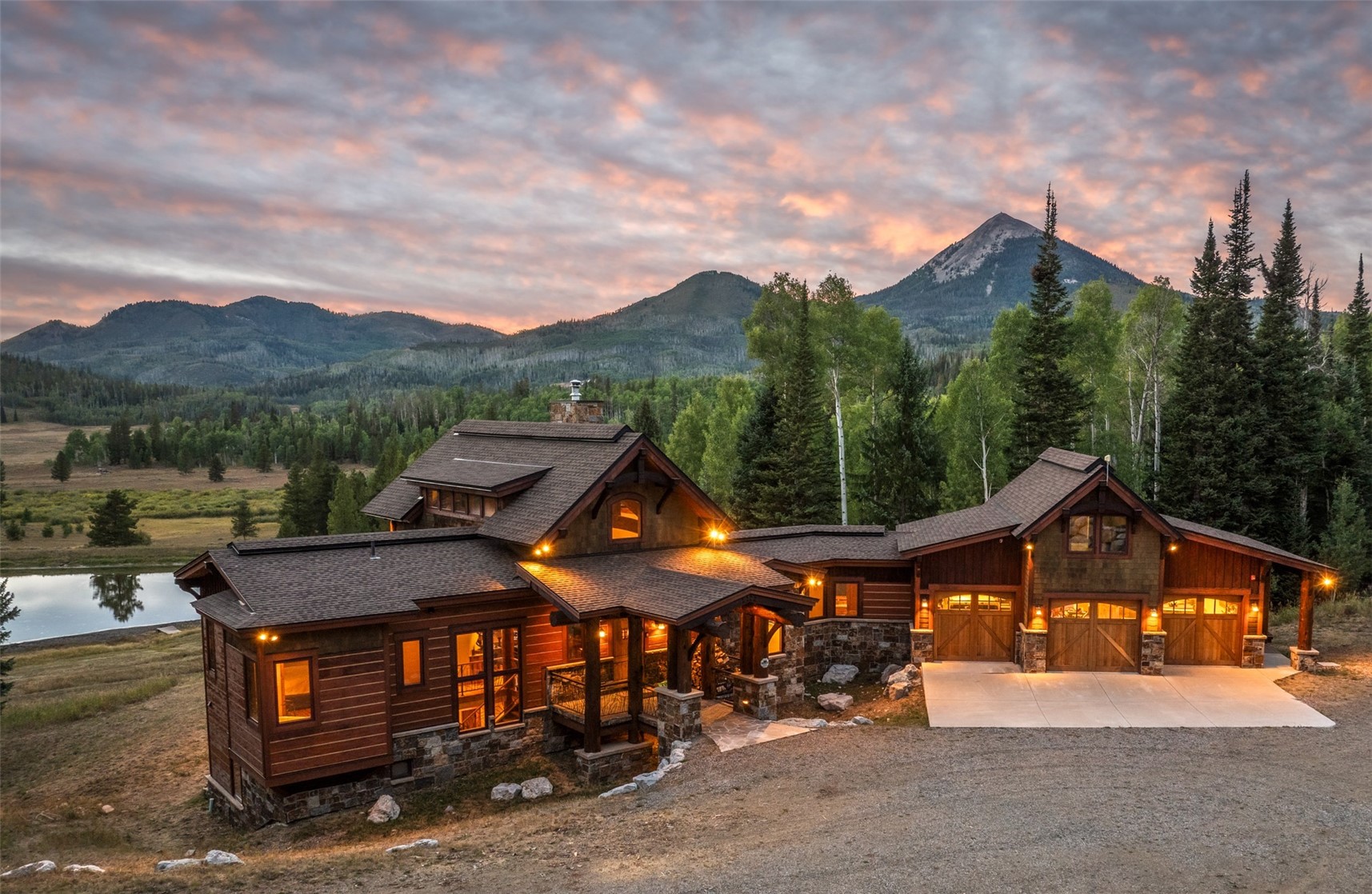 Rustic mountain home glowing at dusk, with a pond and forested mountains behind it.