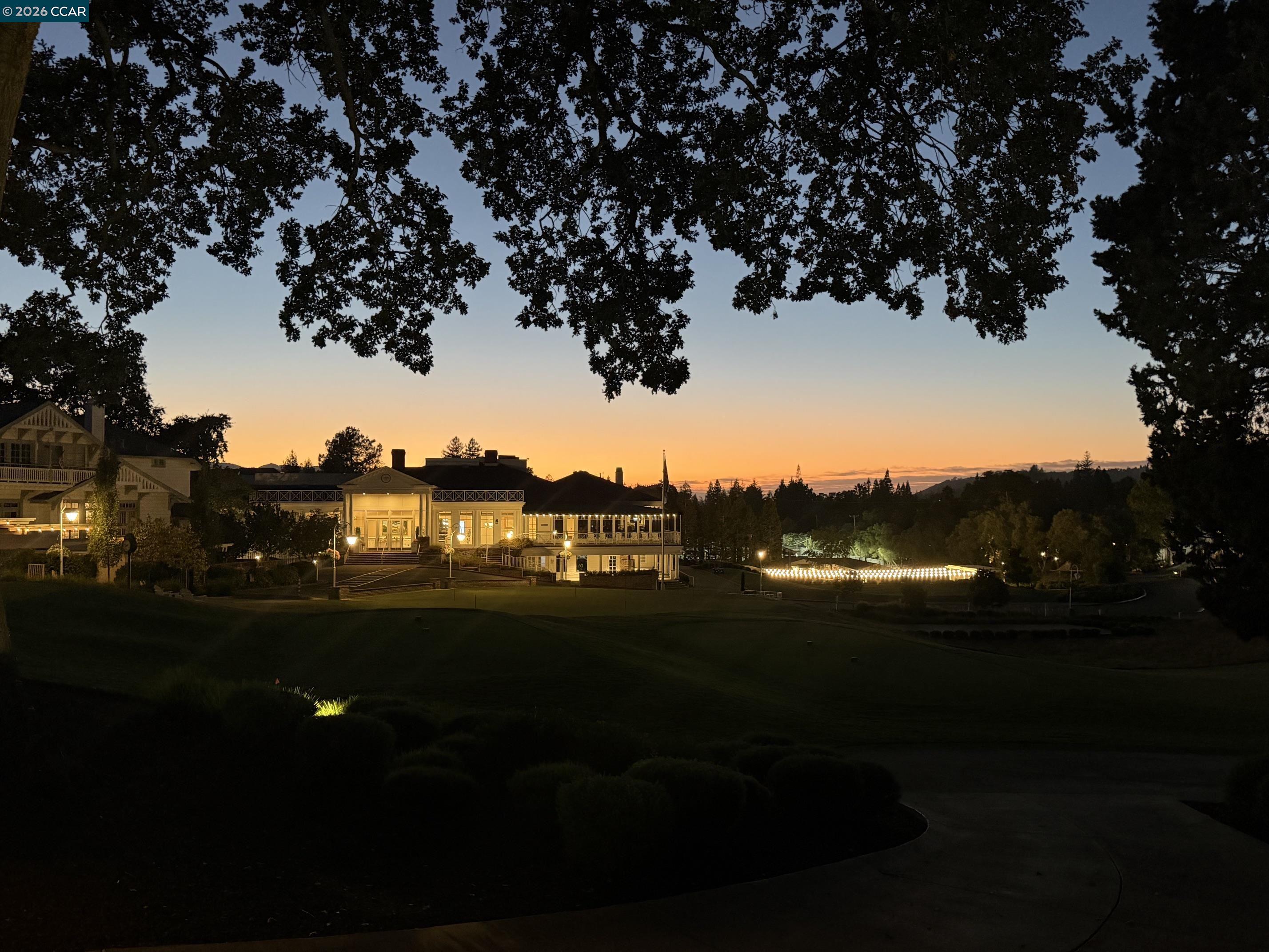 Illuminated clubhouse and broad lawn beneath spreading trees at sunset.