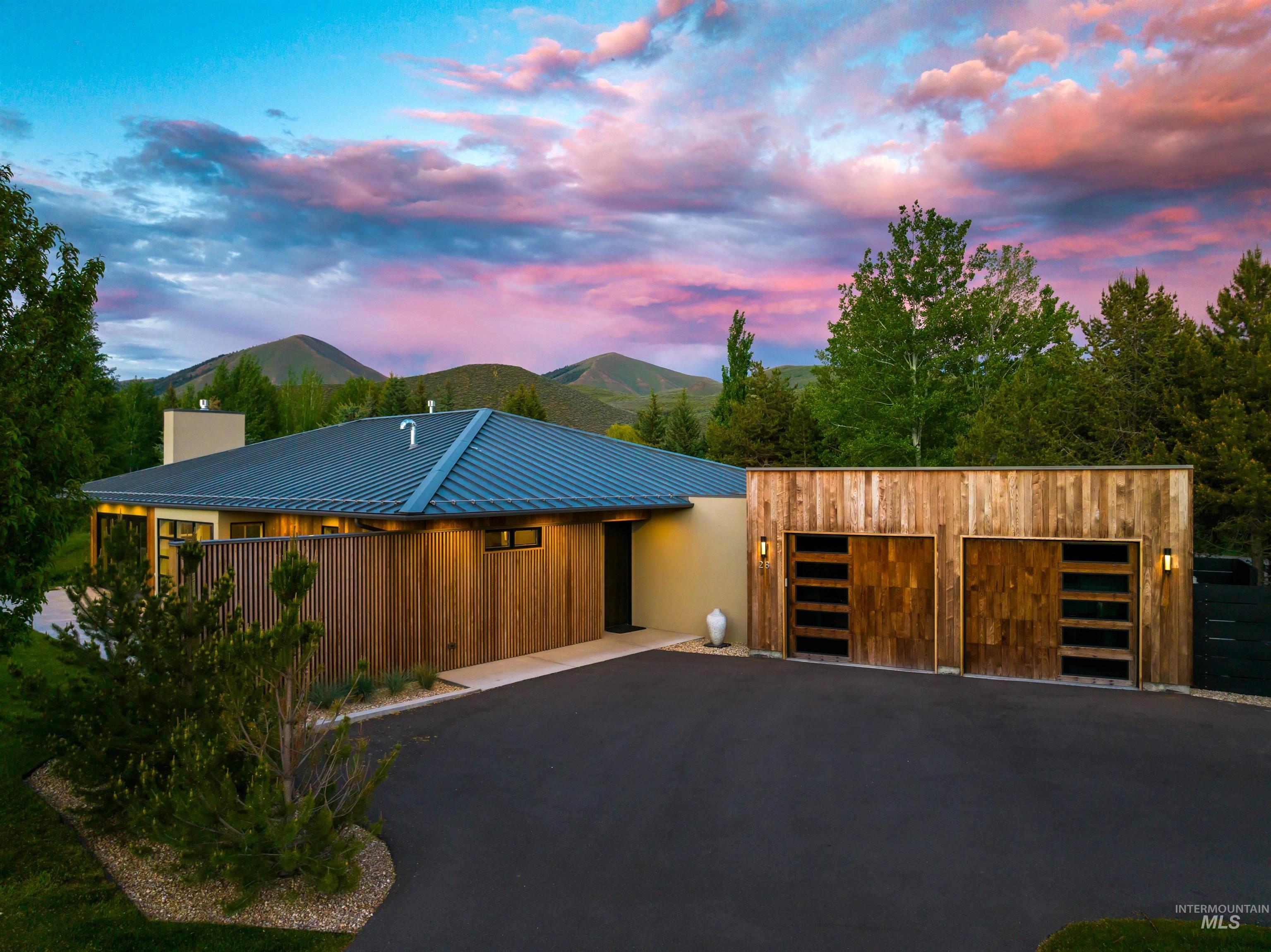 Modern home with wood-clad garage doors and a blue metal roof, set against foothills at dusk.
