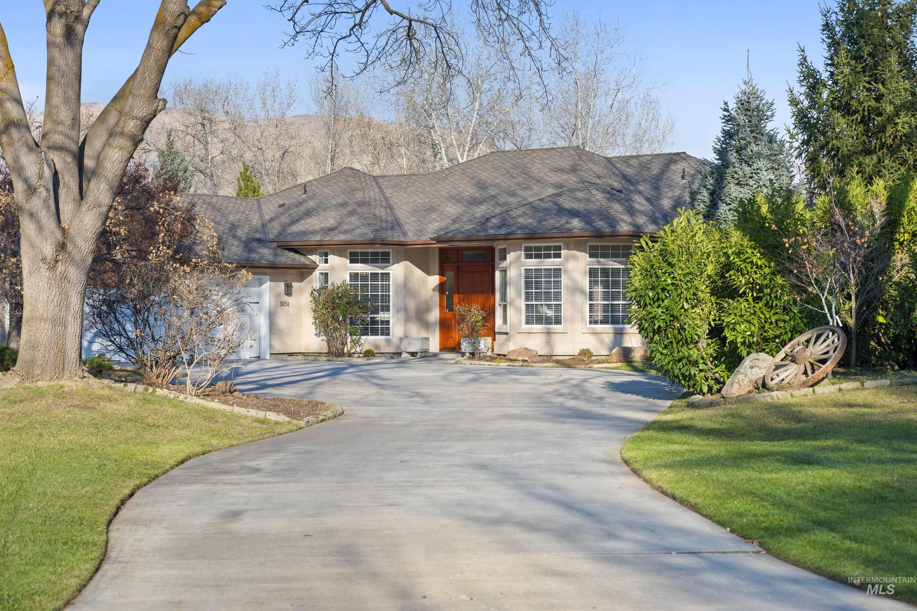Single-story stucco home with a reddish wood entry, curved driveway, mature trees, and landscaped lawn.