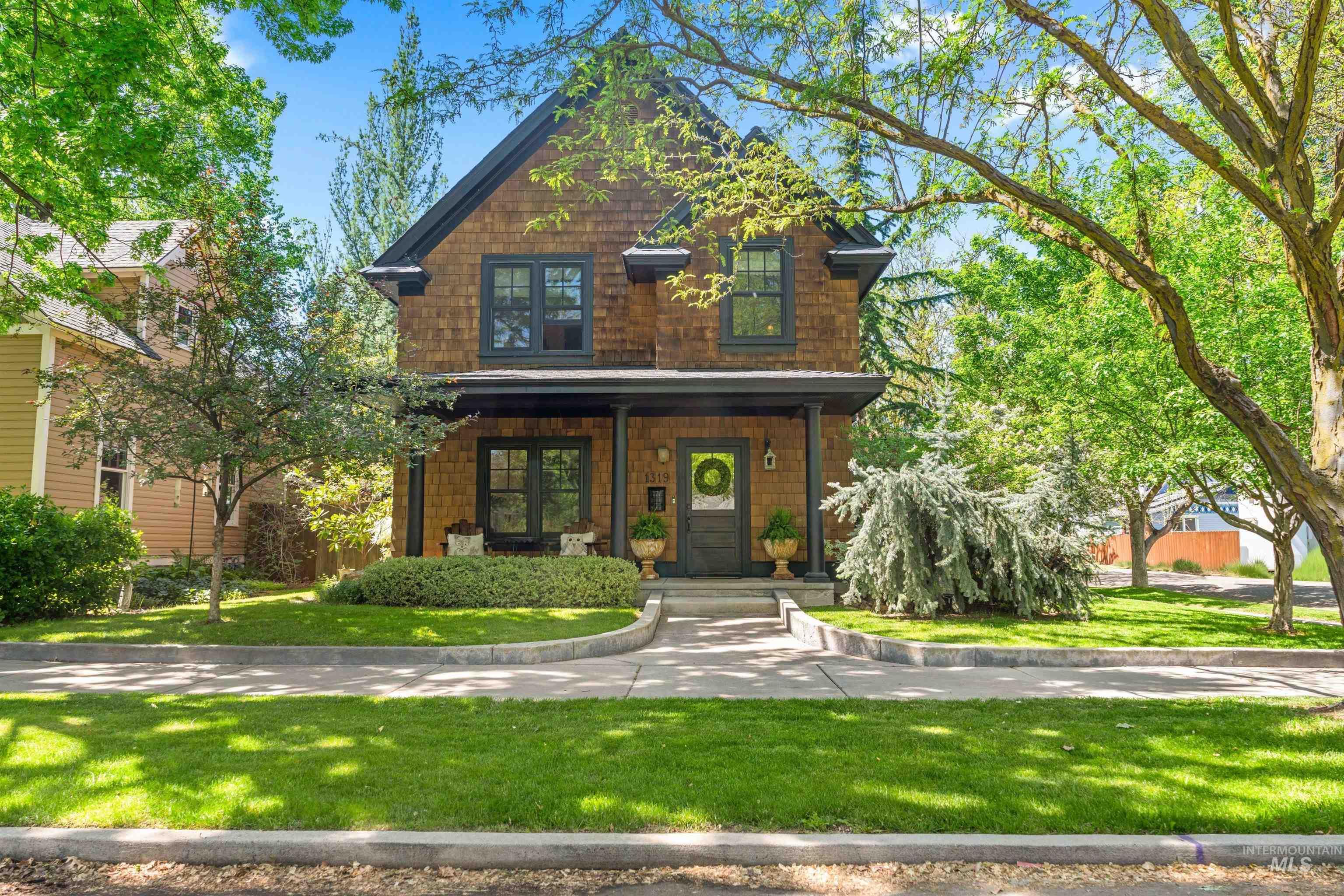 Two-story brown shingle home with a covered porch, leafy trees, and a landscaped front yard.