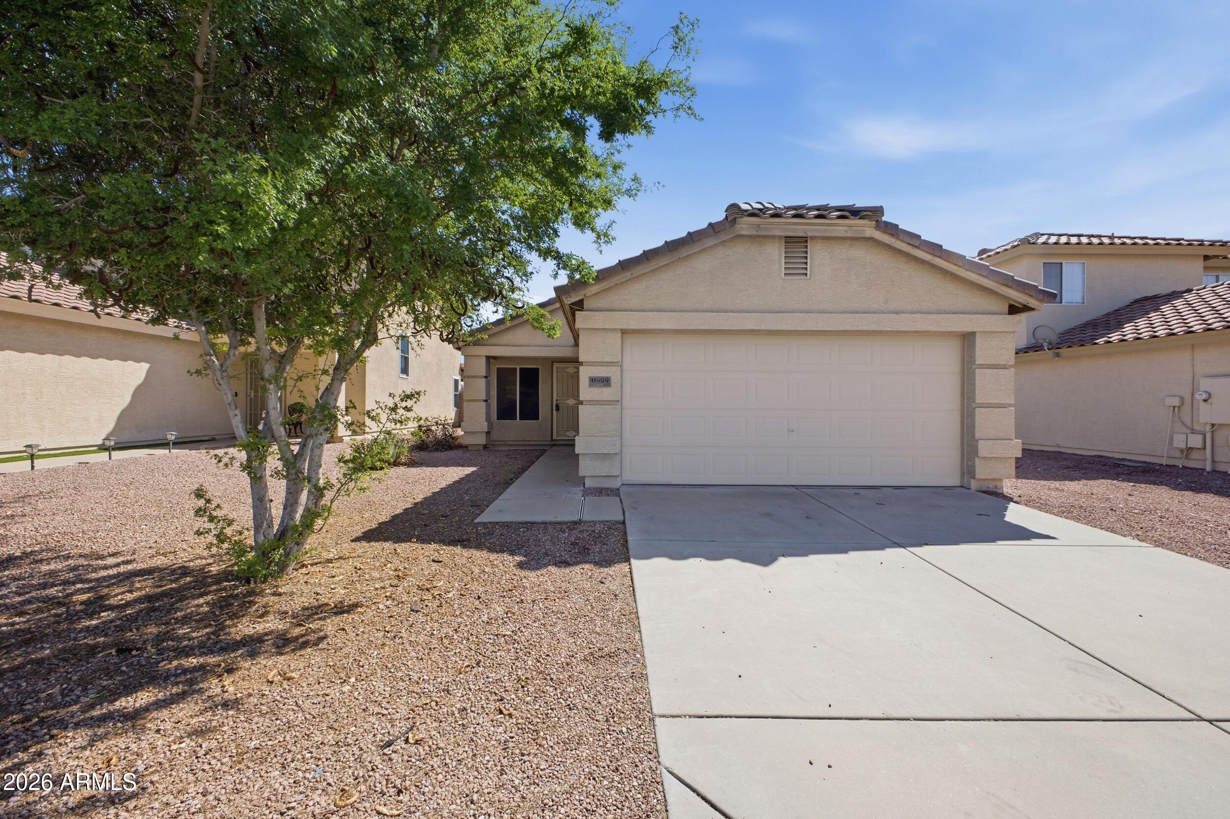 Beige single-story home with a two-car garage, wide driveway, gravel landscaping, and a large shade tree.