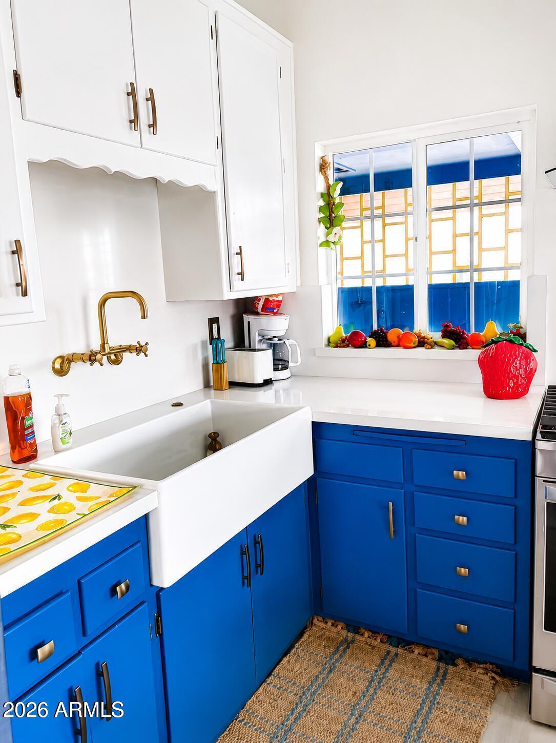 Kitchen with blue cabinets, a white farmhouse sink, brass faucet, and fruit-lined window.