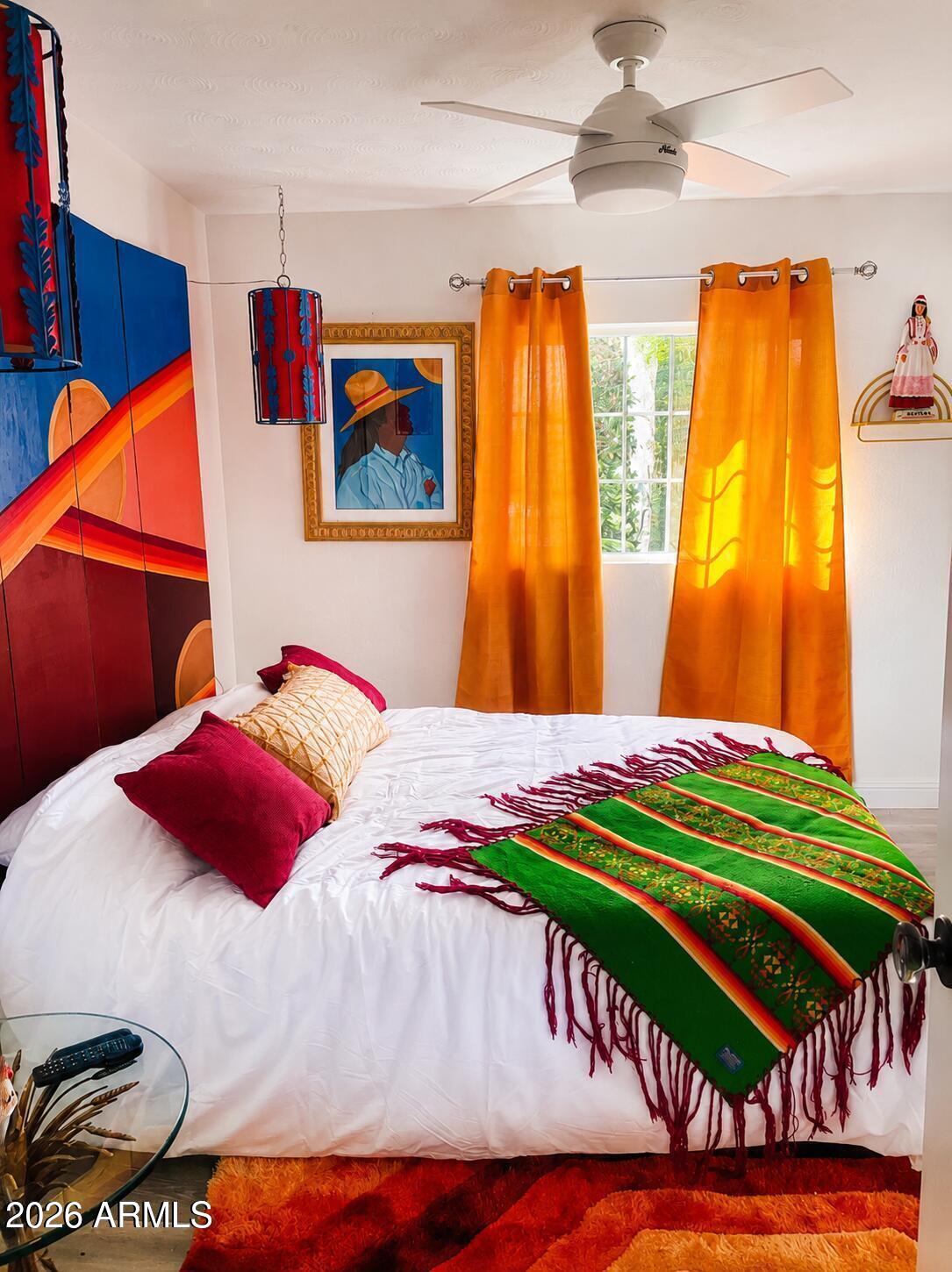Bedroom with a white bed, vivid green throw, orange curtains, and bold blue and red wall art.
