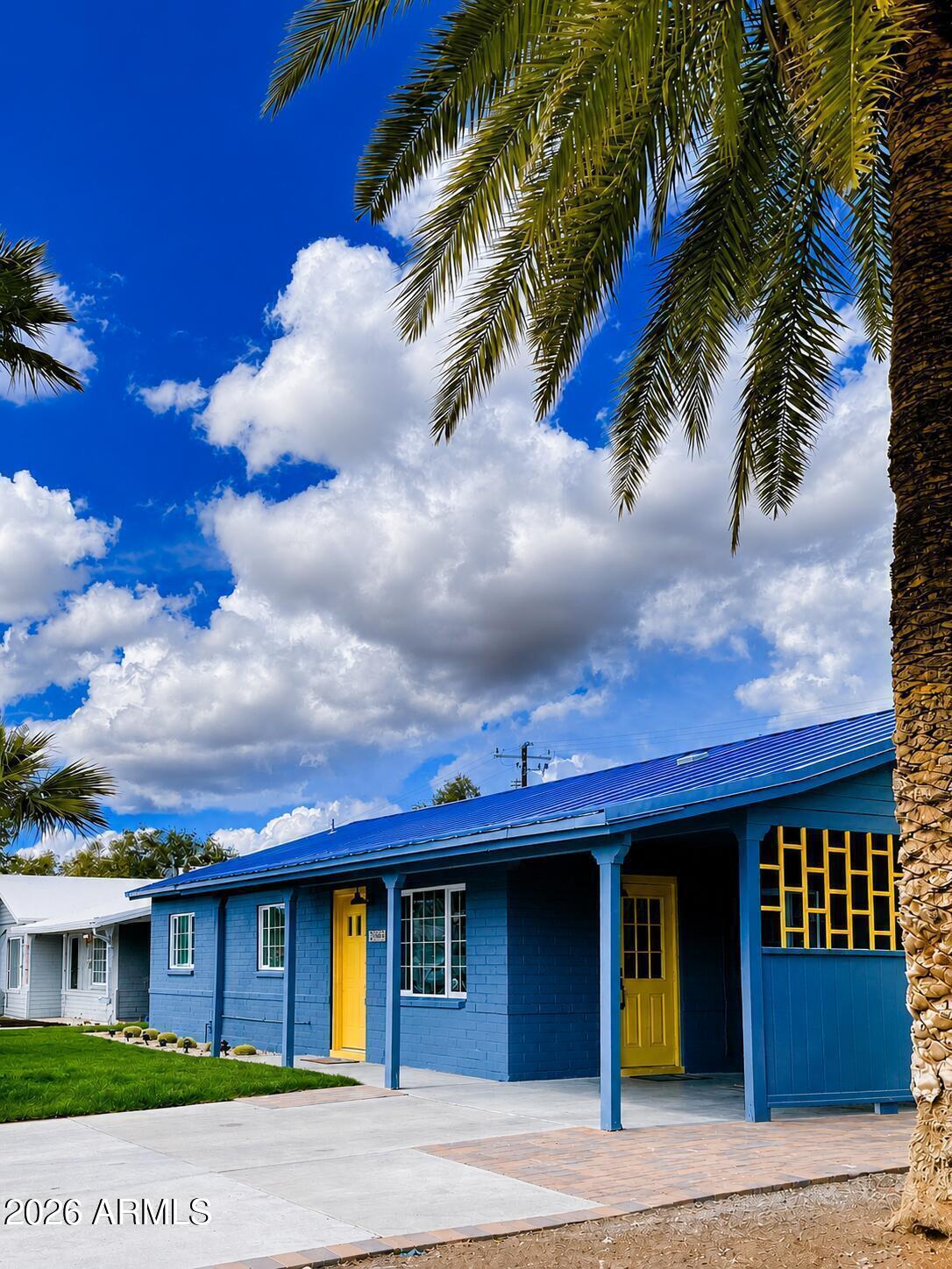 Blue single-story house with yellow doors and trim, framed by palm trees beneath a cloud-filled sky.