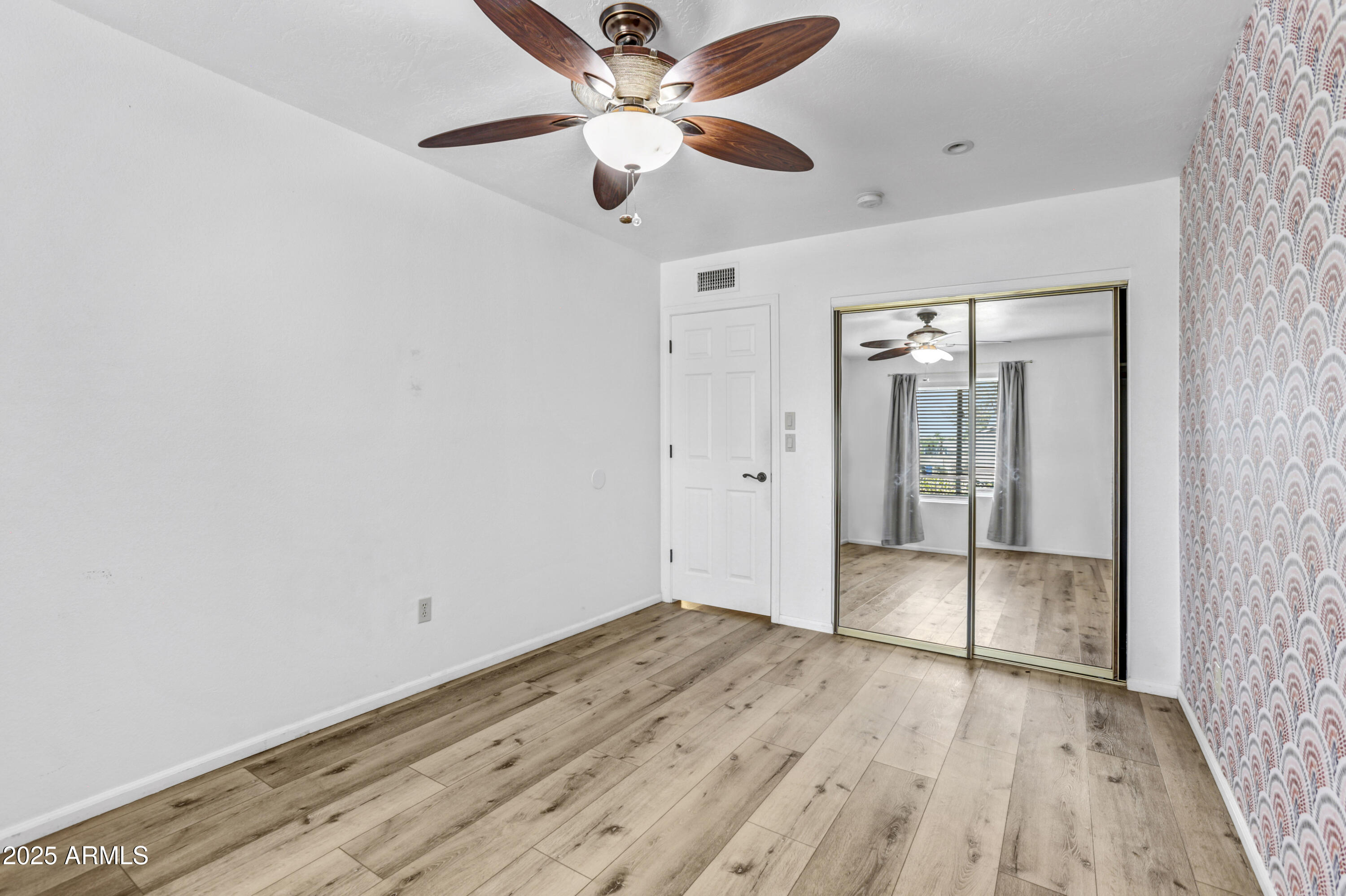 Empty bedroom with mirrored closet doors, wood-look flooring, and patterned wallpaper.