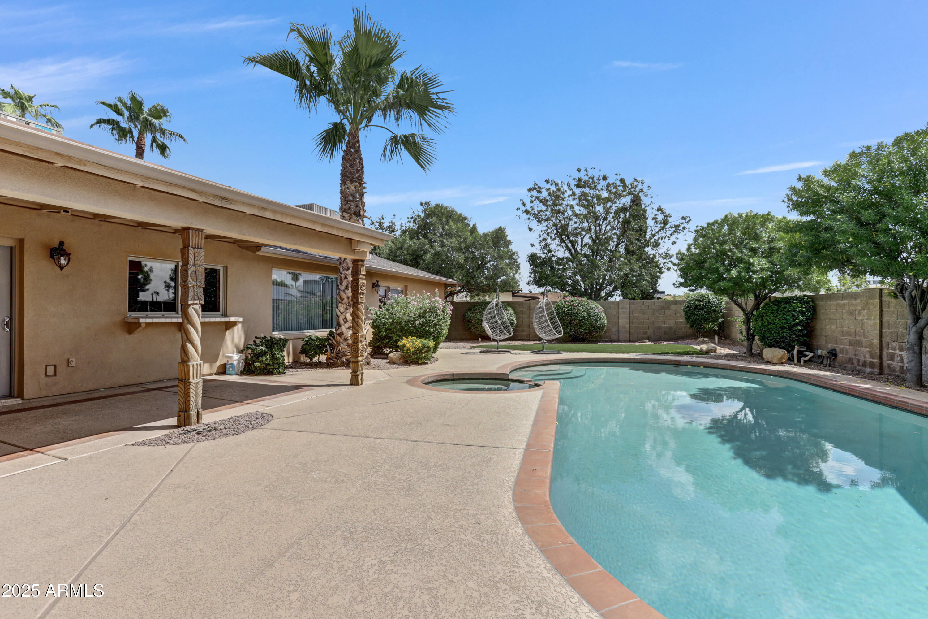 Backyard pool and spa beside a broad patio, palm trees, and two hanging chairs.