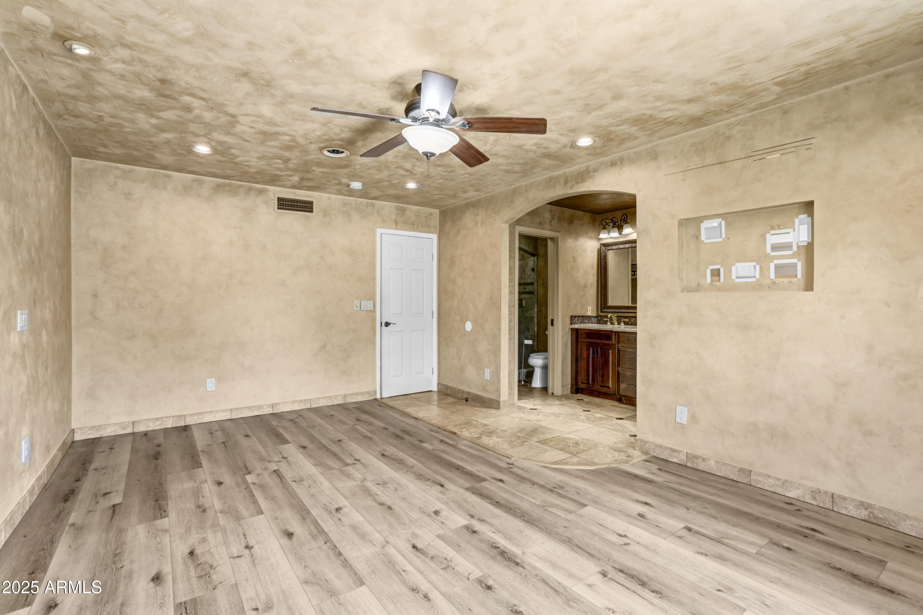 Empty bedroom with wood-look flooring, ceiling fan, and an arched opening to an ensuite bathroom.