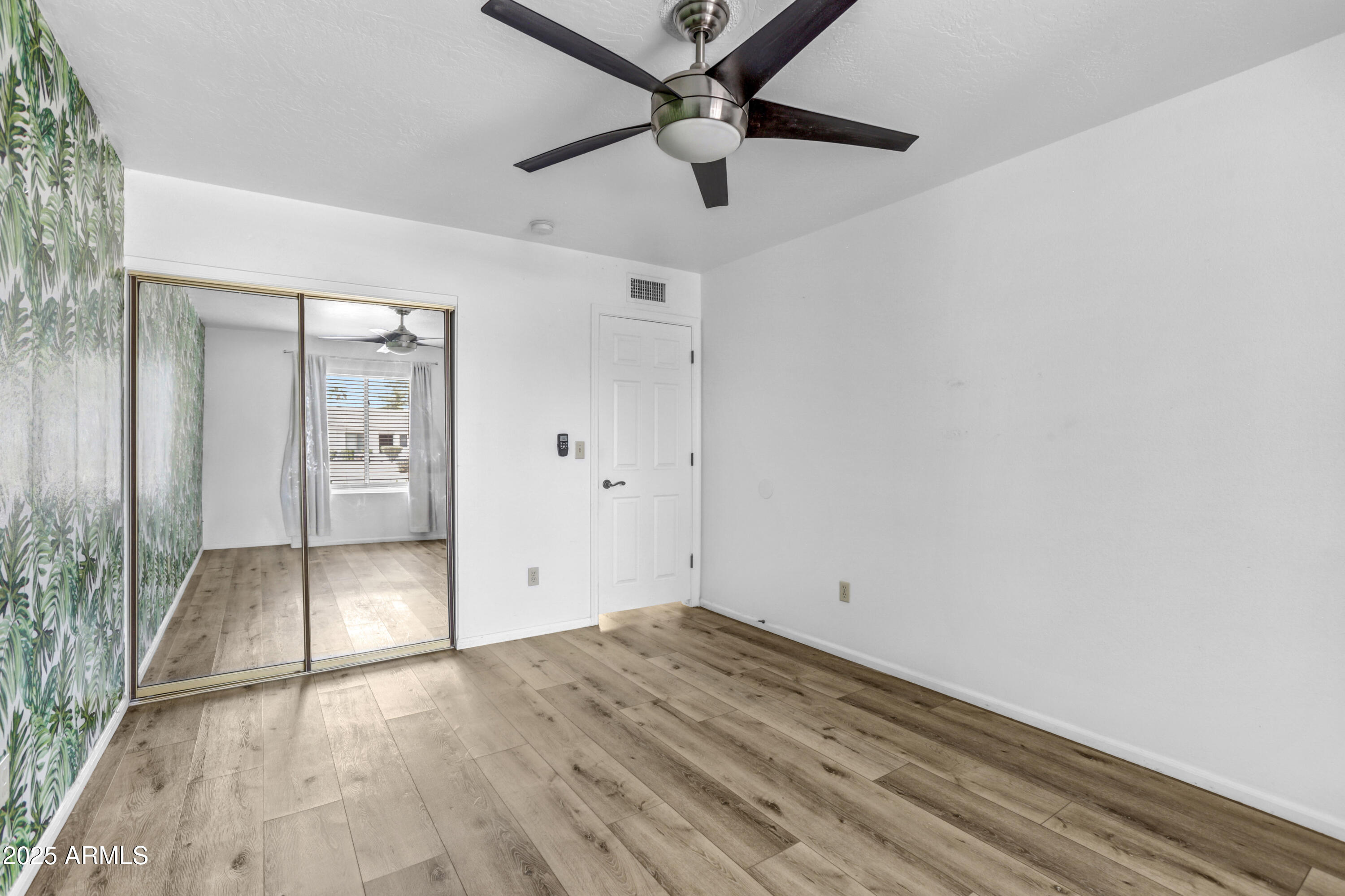 Empty bedroom with leafy wallpaper, mirrored closet doors, wood-look floors, and a dark ceiling fan.
