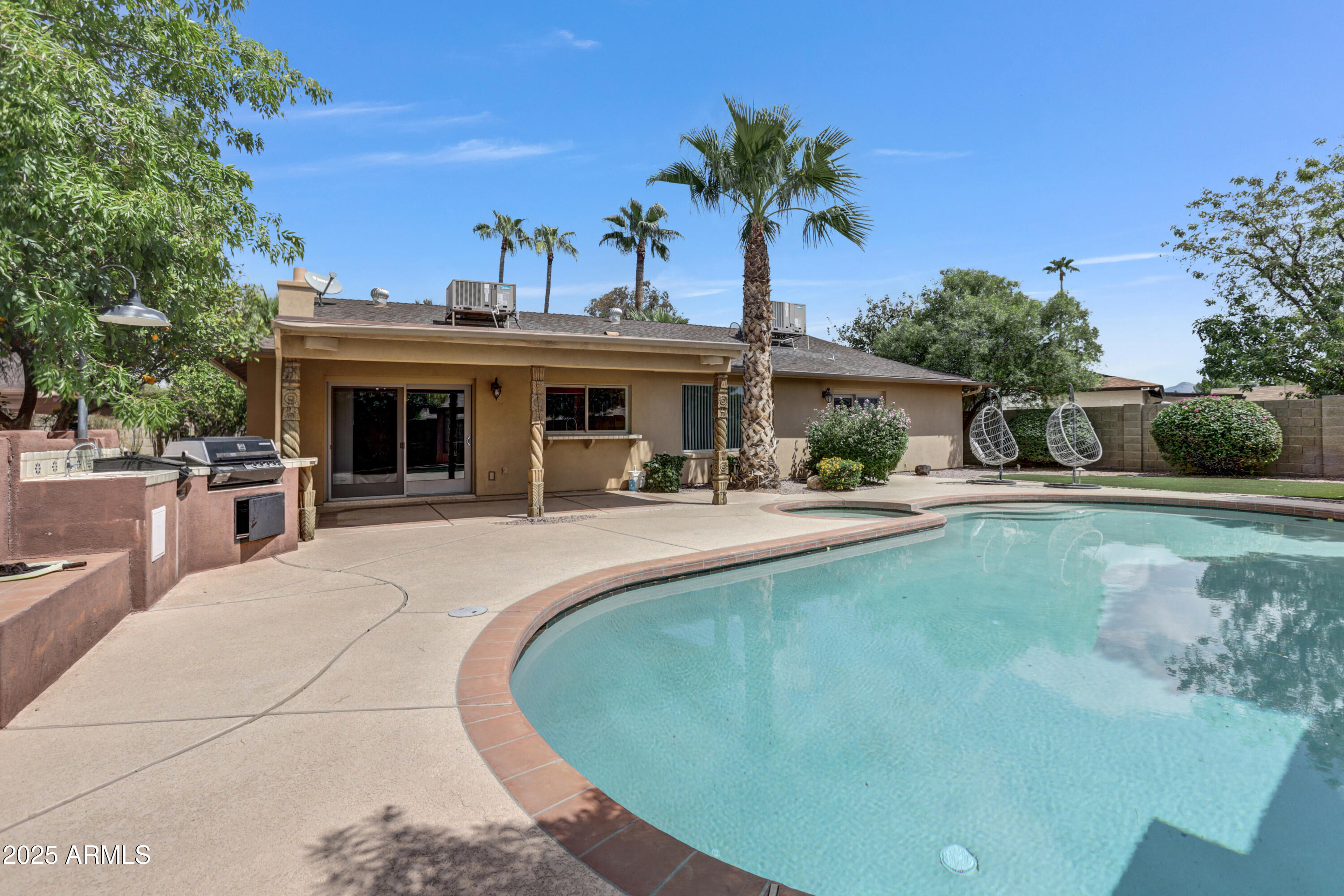 Back of a single-story home beside a curved pool, with a built-in grill and hanging chairs.